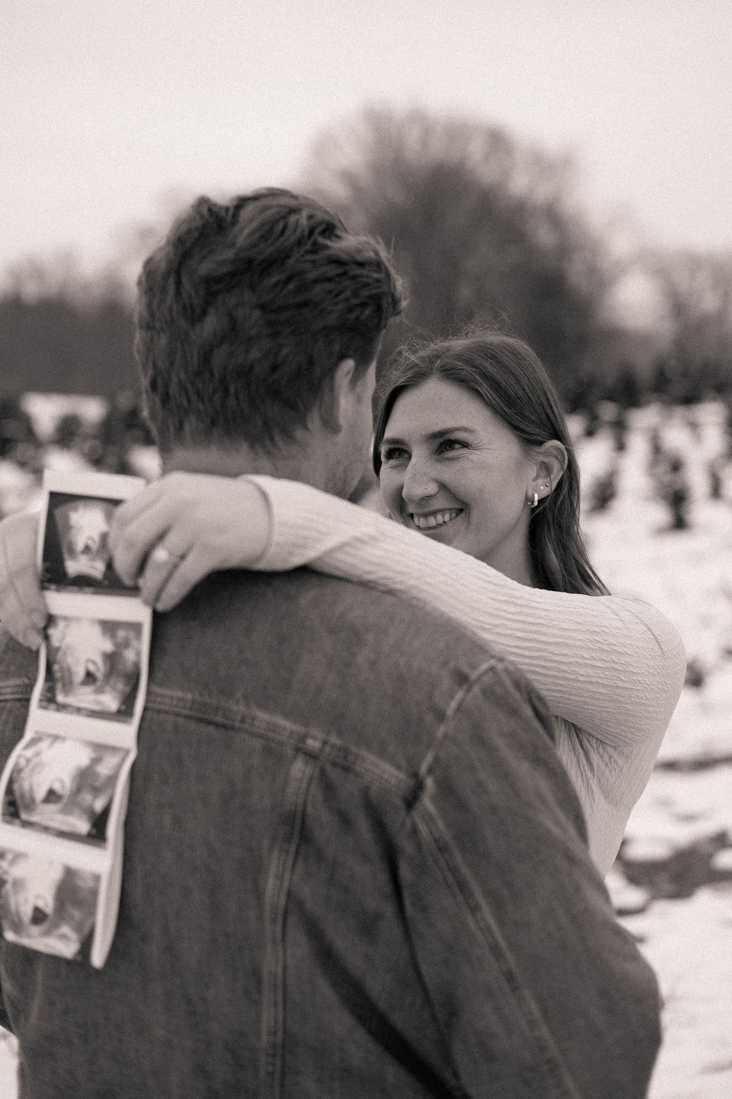Black and white photo of smiling woman hugging man holding ultrasound photos in snowy outdoor setting.