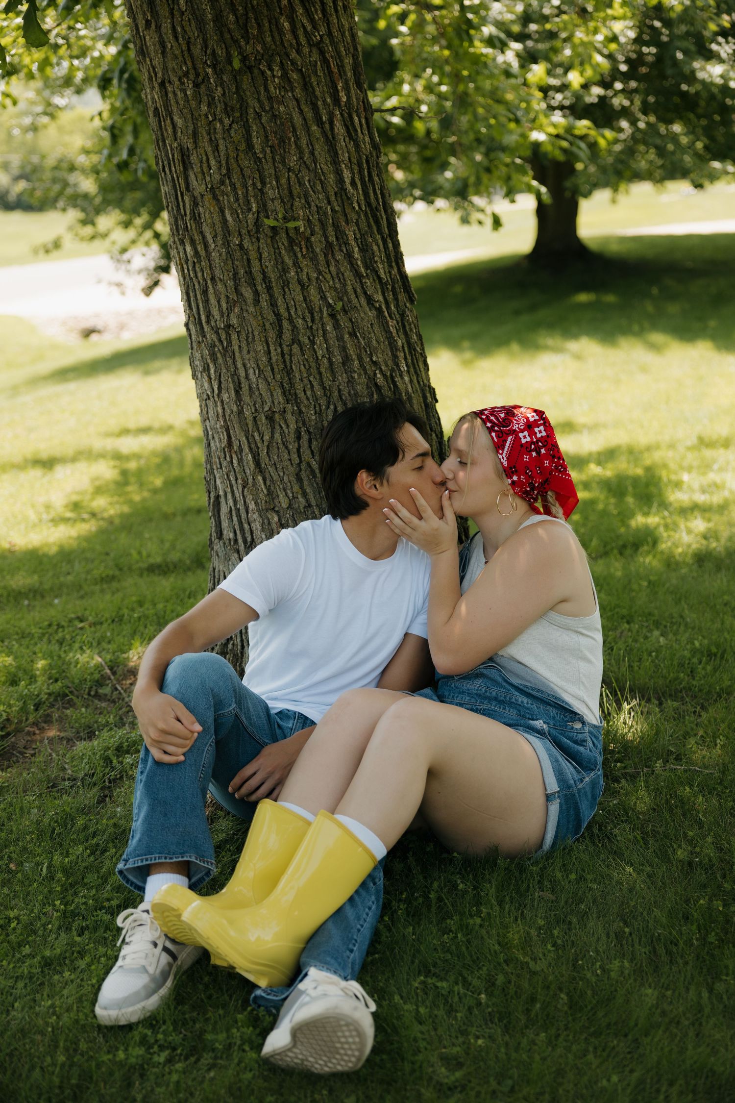 Couple kissing under a tree on green grass, woman in yellow rain boots and red bandana, man in white tee and jeans.