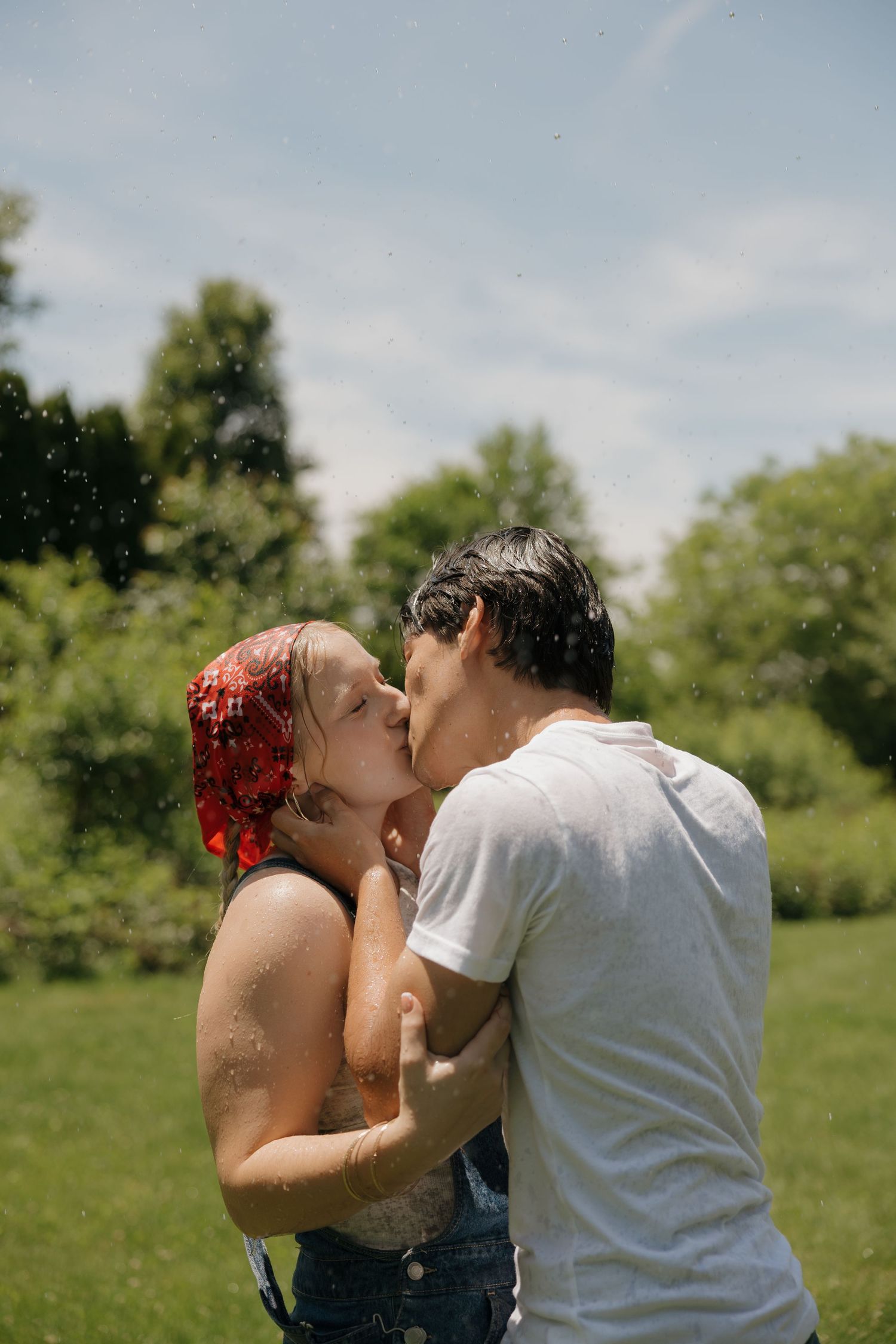 A couple shares a romantic kiss outdoors in a lush green park on a sunny day, with water droplets surrounding them.