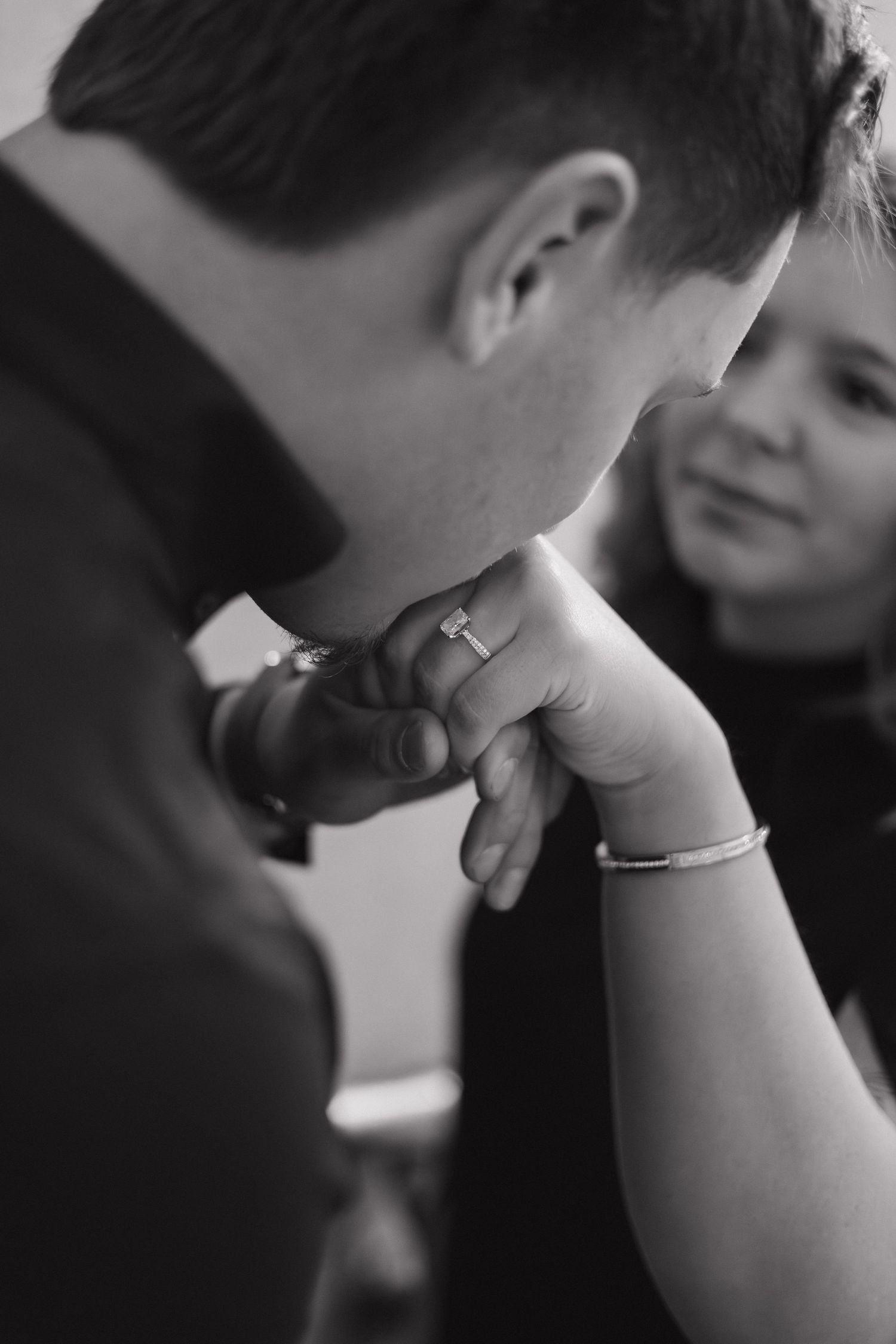 Black and white photo of a man kissing a woman's hand, showcasing her engagement ring and bracelet.