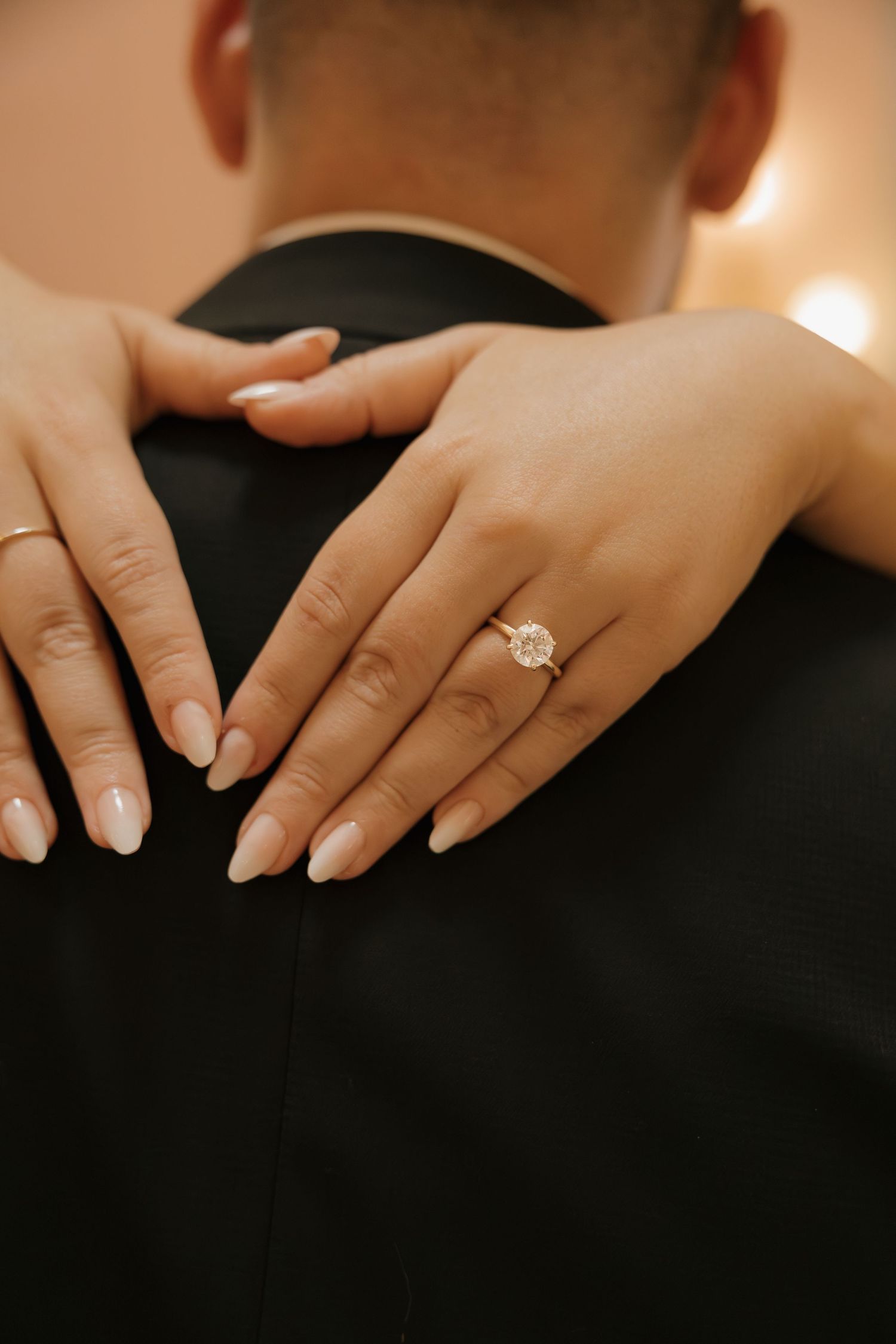 Close-up of womans hand with oval diamond engagement ring resting on mans dark suit jacket.