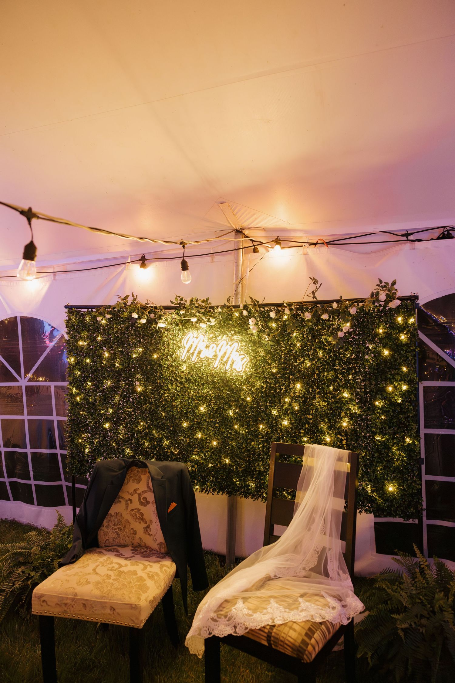 Mr and Mrs neon sign glows on greenery wall backdrop with string lights inside wedding tent.