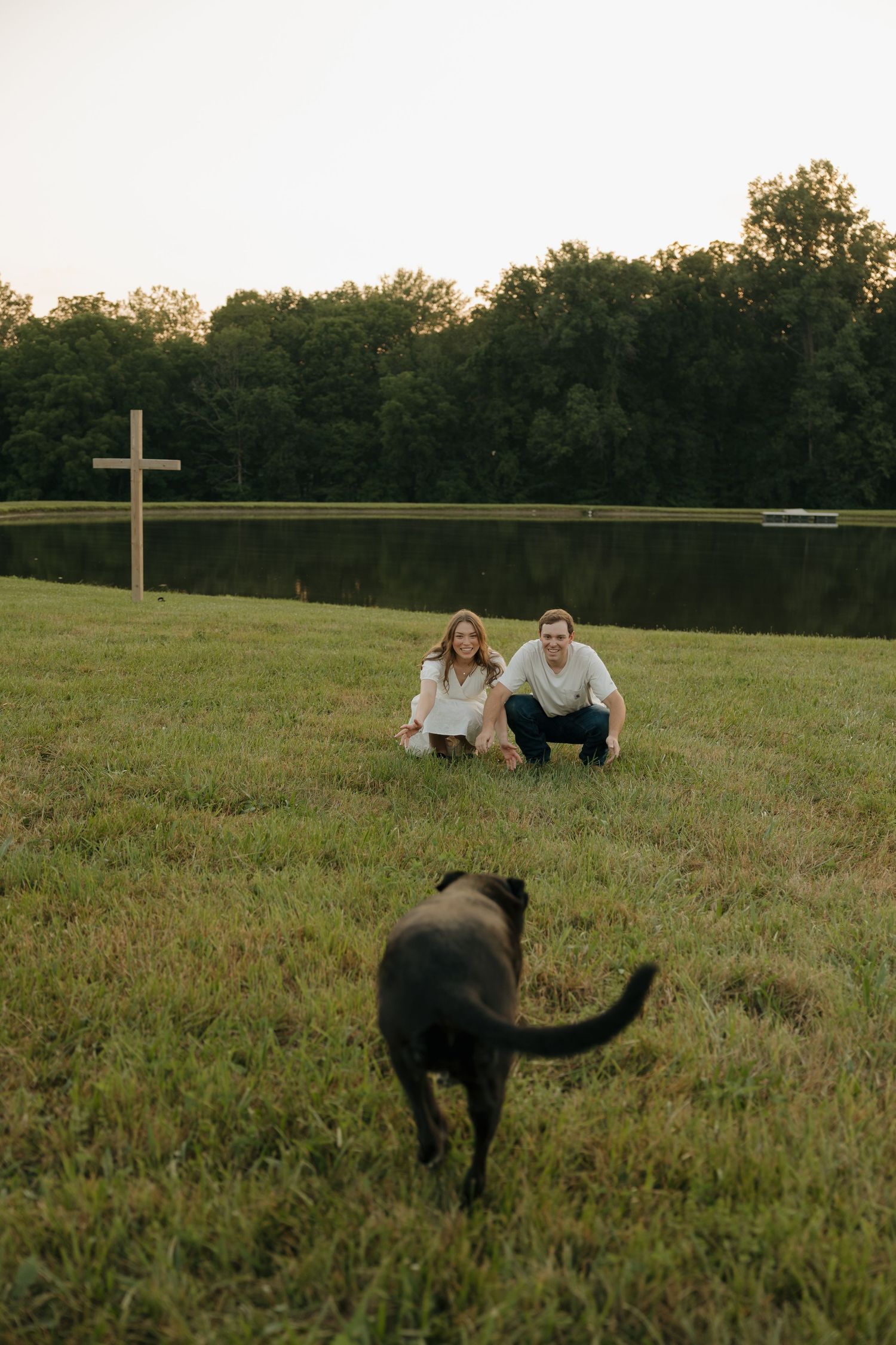 Couple crouching on green grass near a pond with a wooden cross, as a black dog approaches them at dusk.