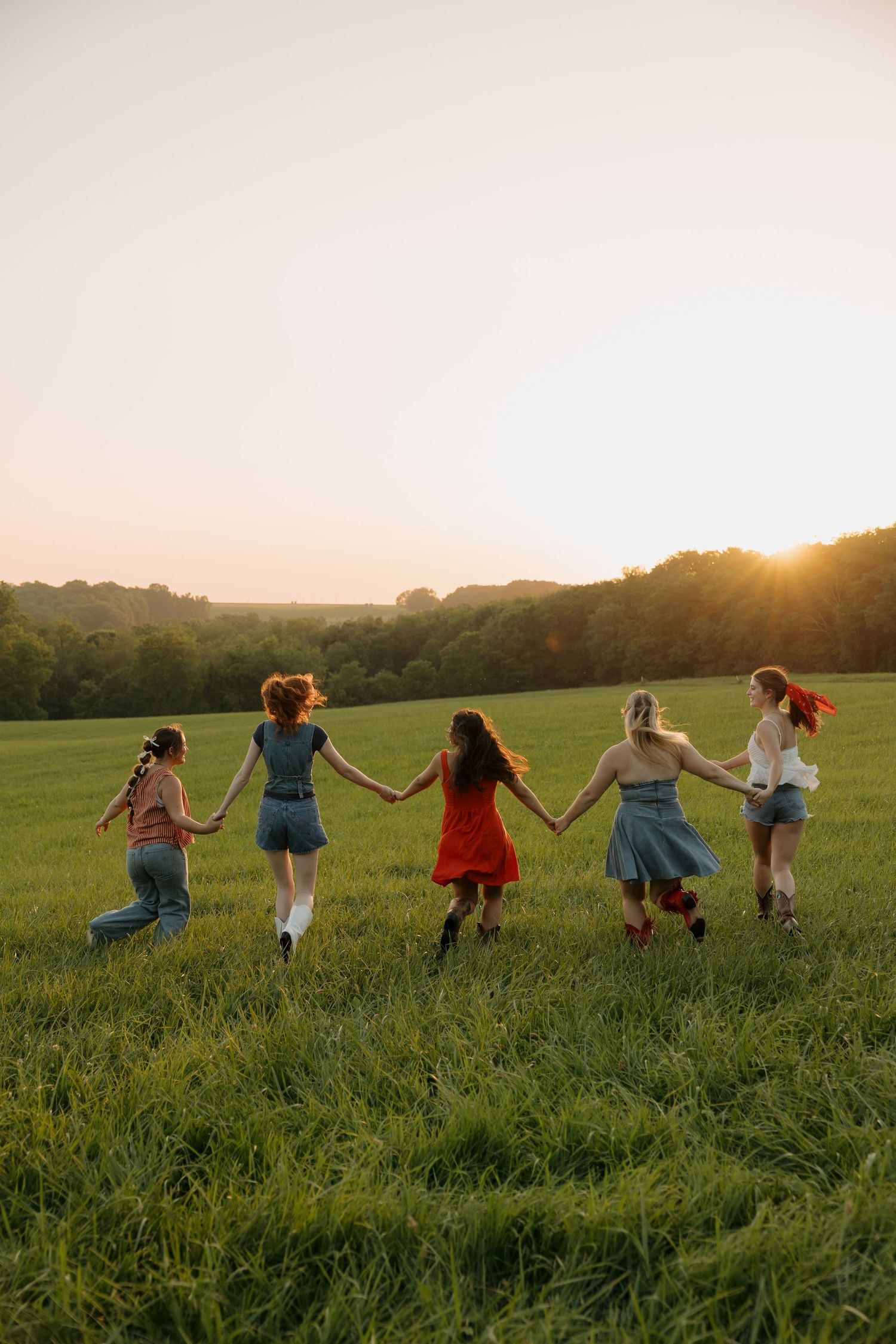 Five young women holding hands run through a green field at golden sunset, wearing colorful summer dresses and casual outfits.