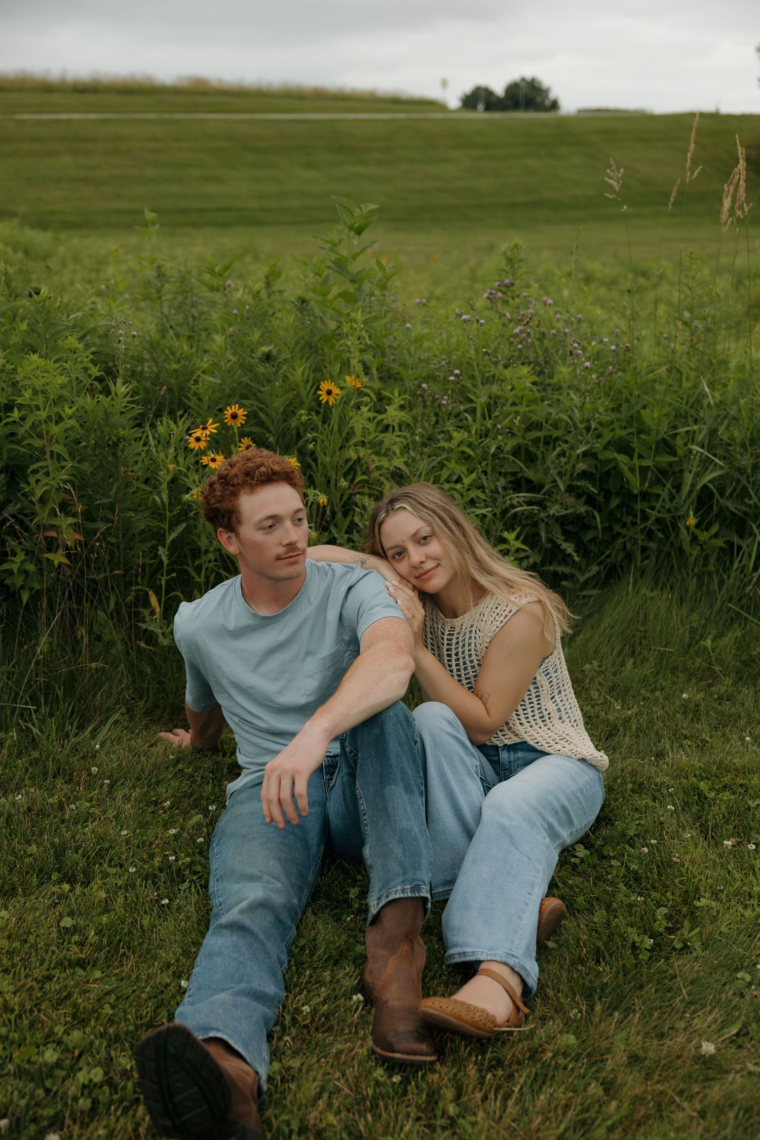 A young couple sitting together in a lush green field, wearing jeans and casual tops, surrounded by wildflowers.