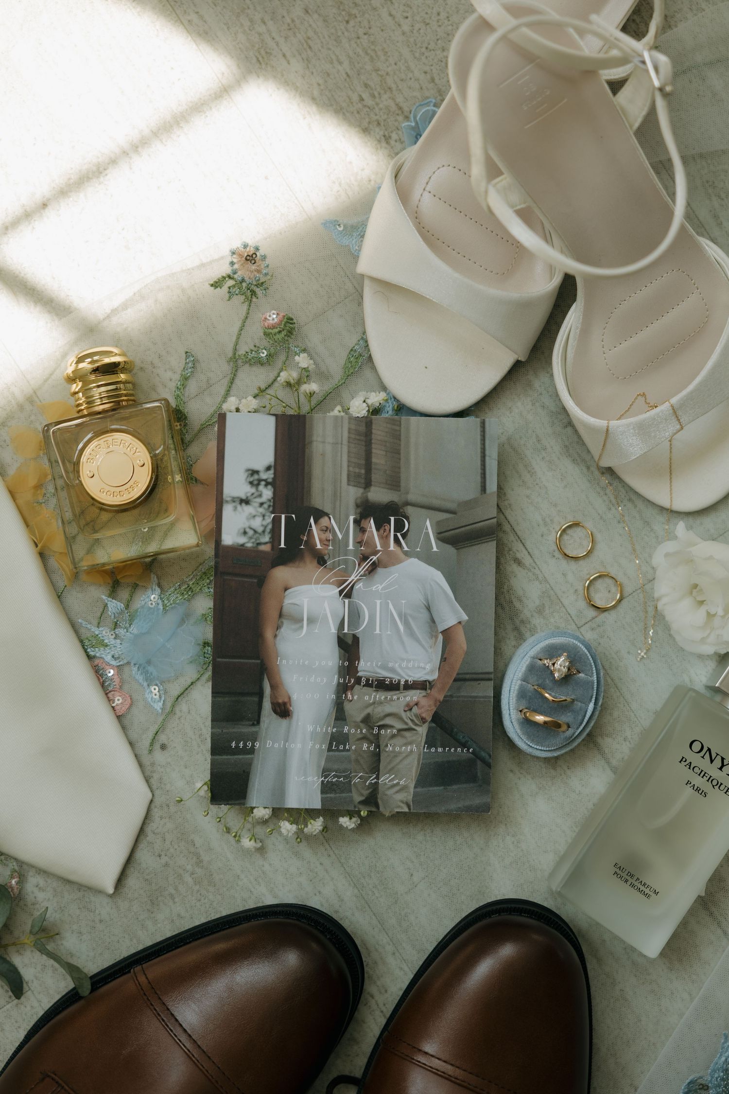 Elegant wedding flat lay with invitation, white heels, gold perfume, rings, and brown dress shoes on floral backdrop.