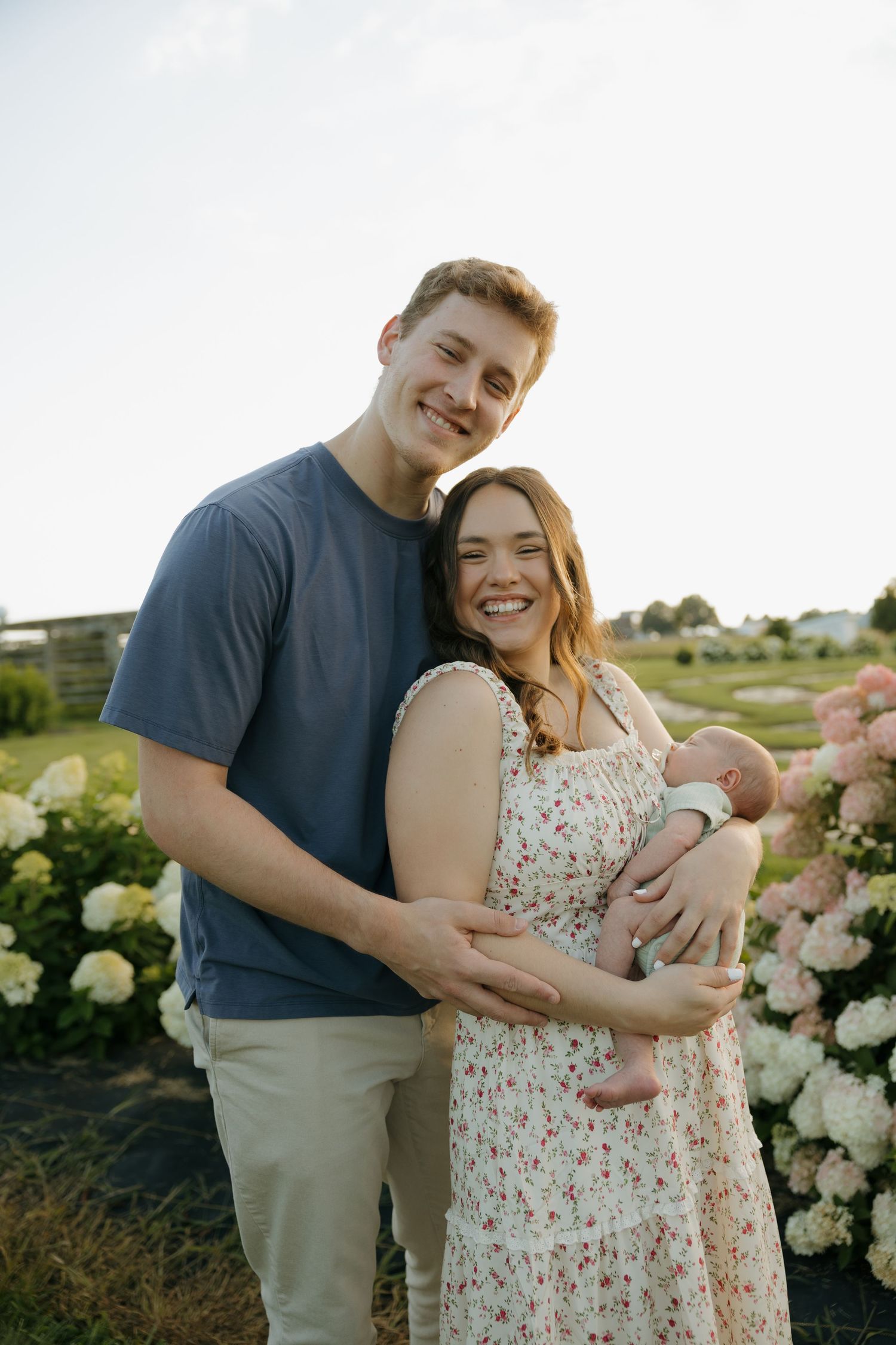 Young couple smiling outdoors holding newborn baby, surrounded by blooming hydrangeas in a sunny garden setting.