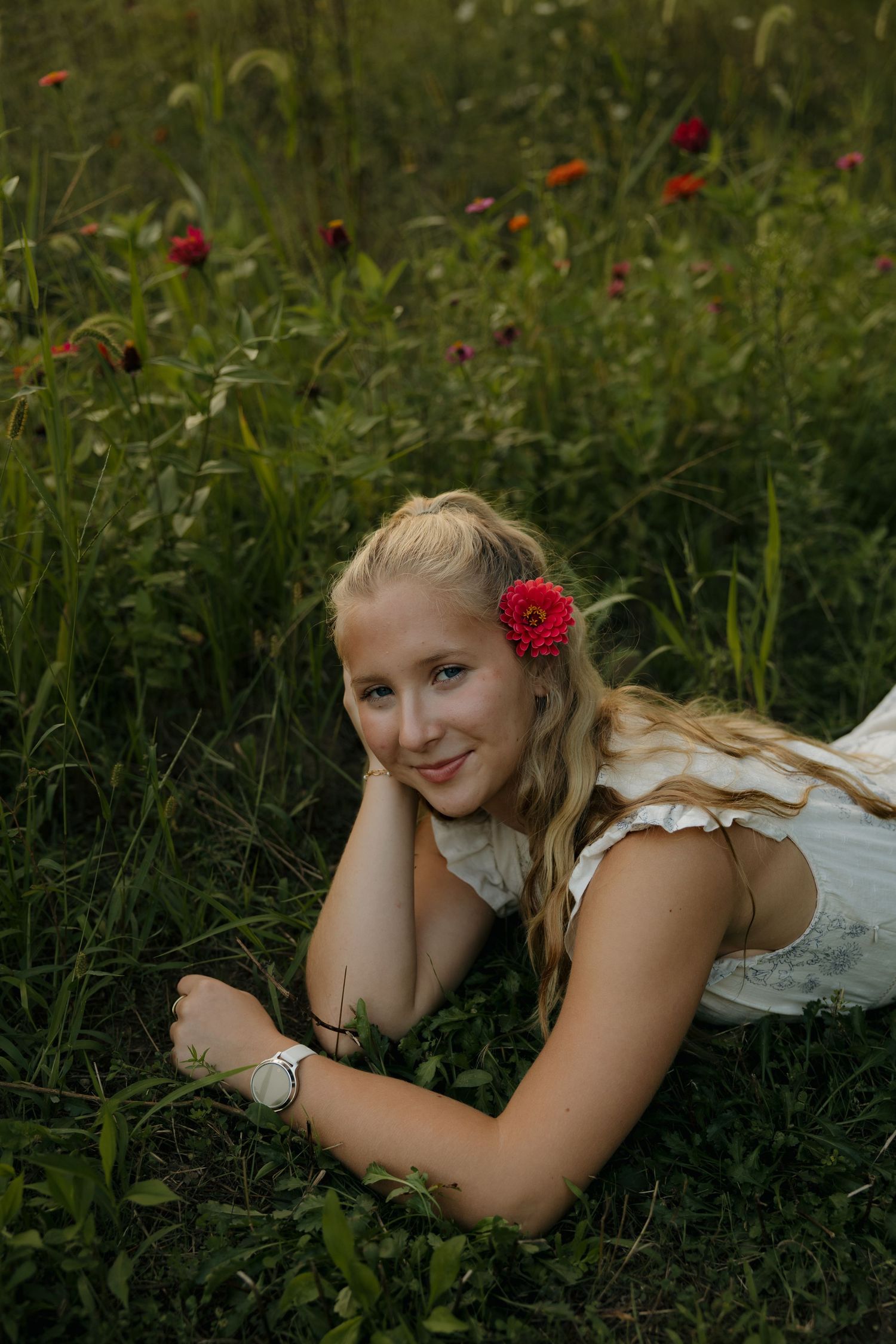 Young blonde woman lying in a wildflower meadow, wearing a white dress with a red flower in her hair.