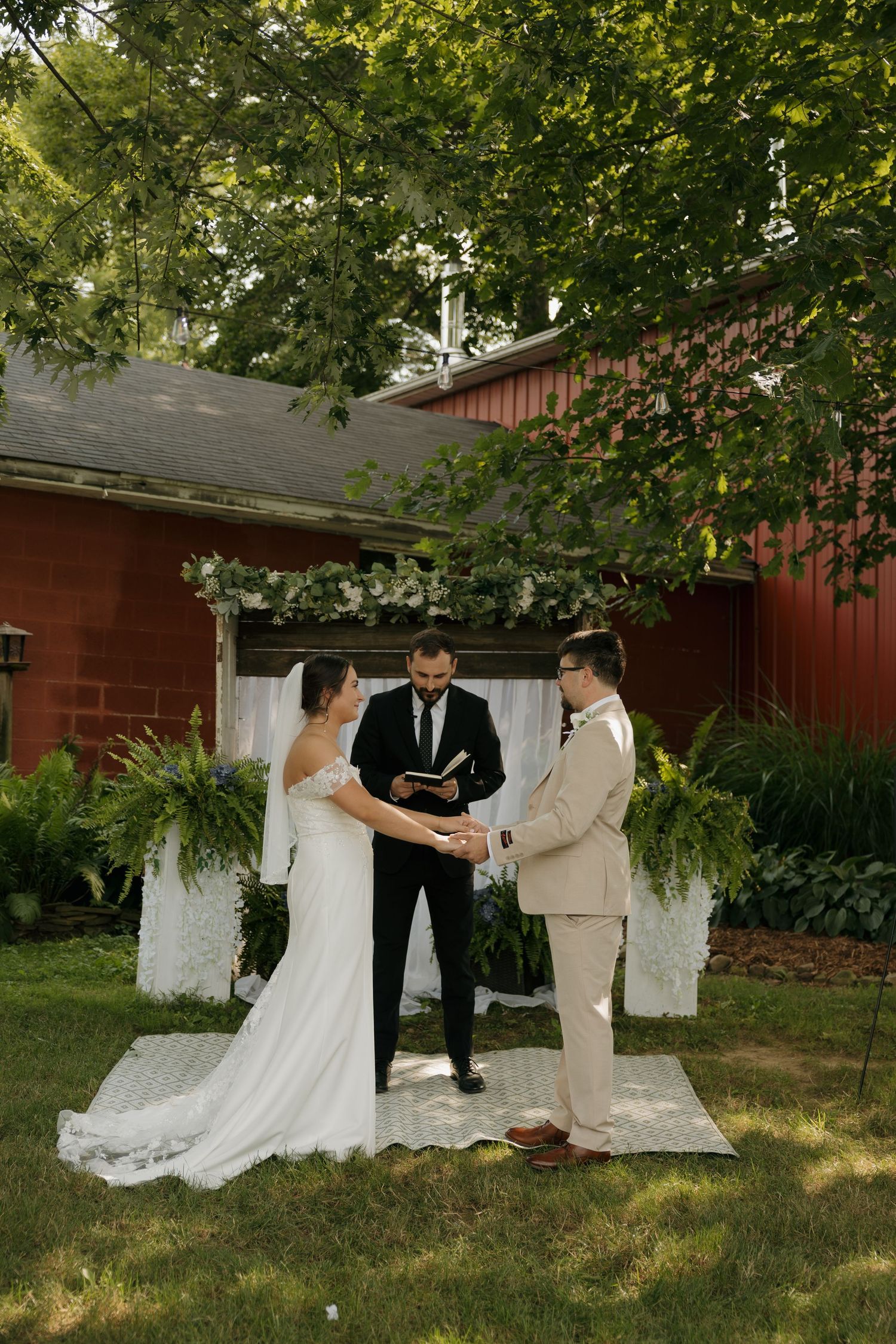 Bride and groom exchange vows at outdoor wedding ceremony beneath a tree with white floral arch and red barn backdrop.