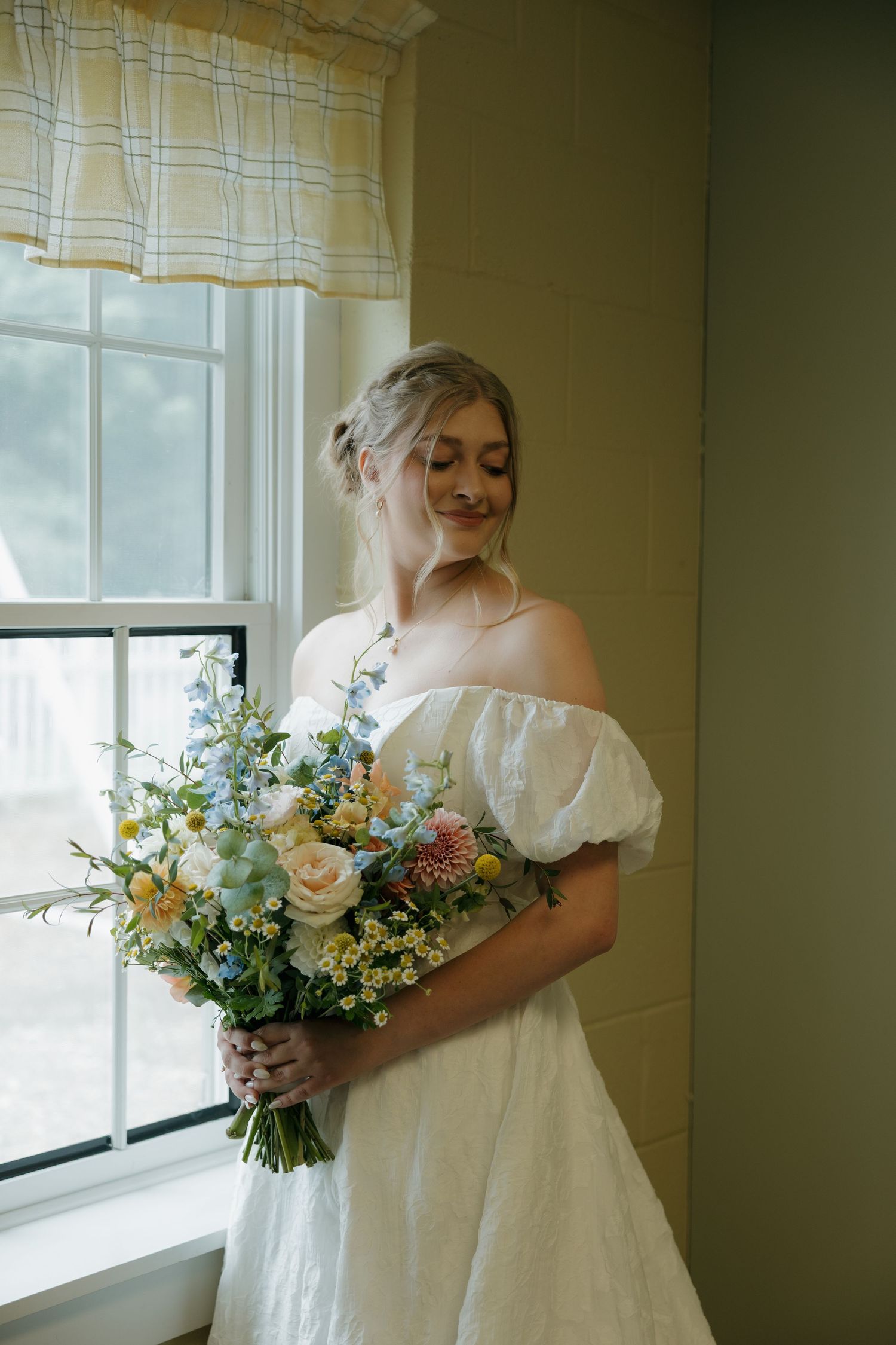Bride in off-shoulder white gown holds colorful spring bouquet by window.