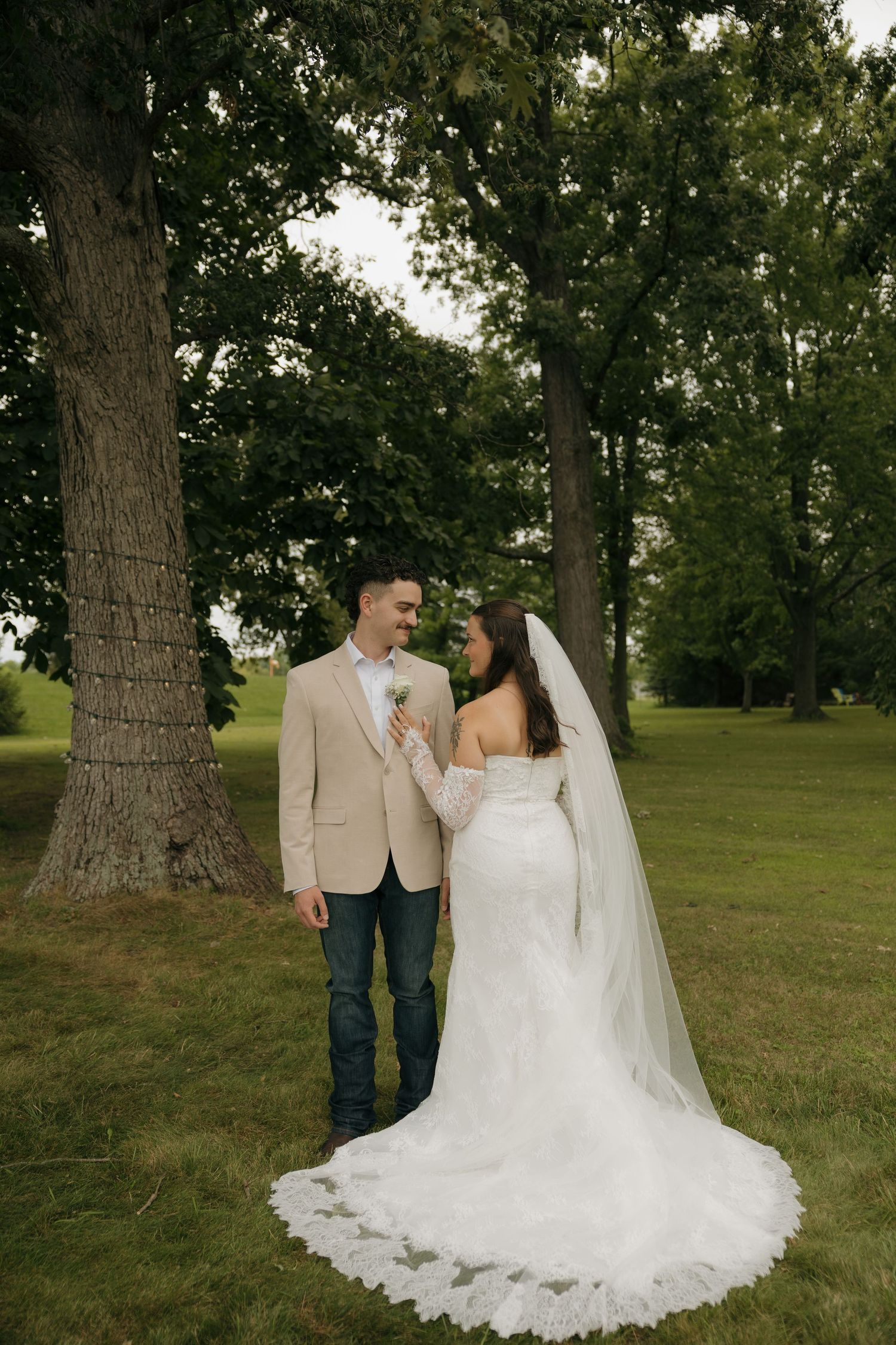Bride in lace gown with veil and groom in tan blazer and jeans share a romantic moment under large trees.