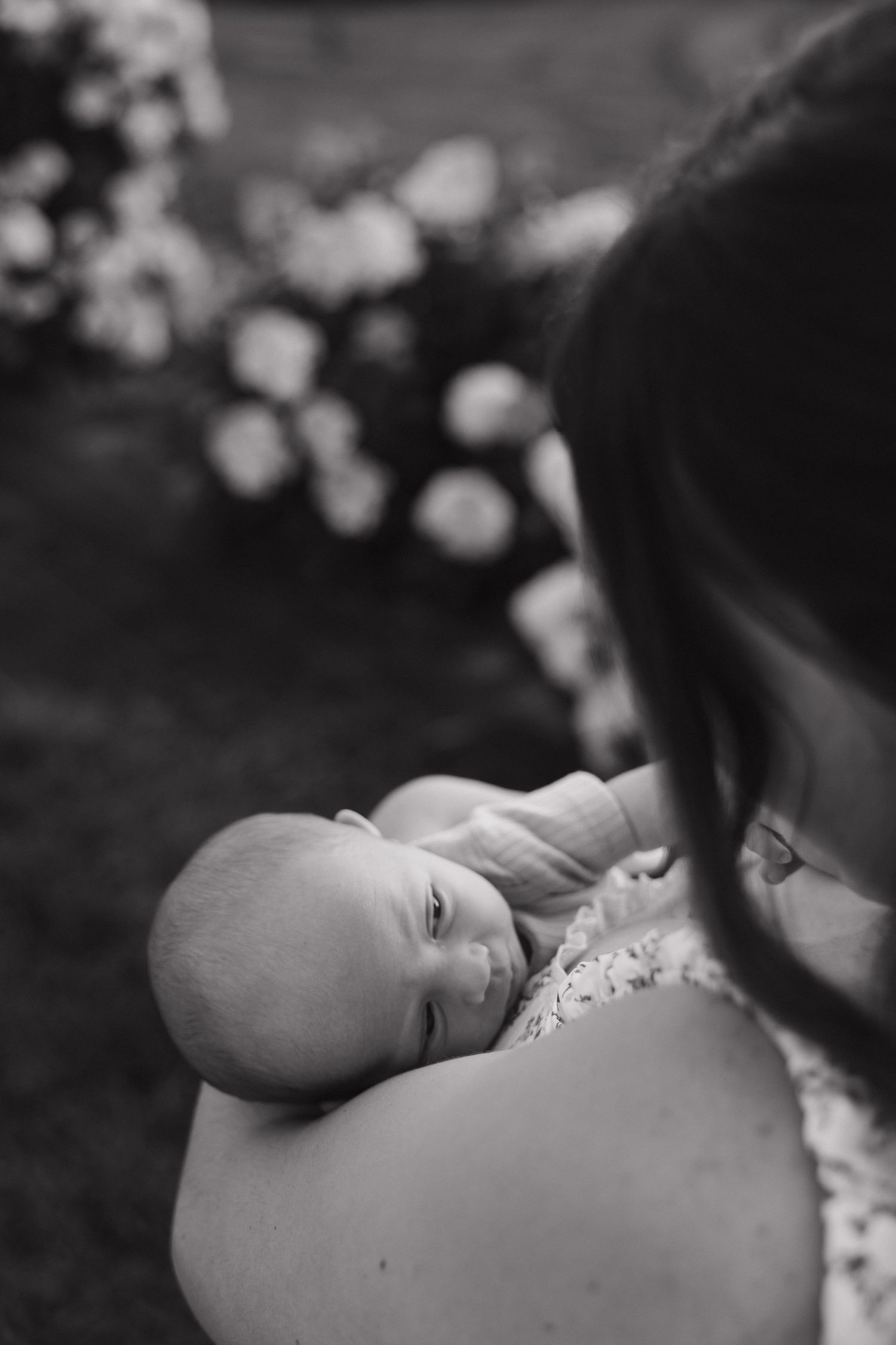 Black and white photo of a mother cradling her newborn baby outdoors with flowers in the background.