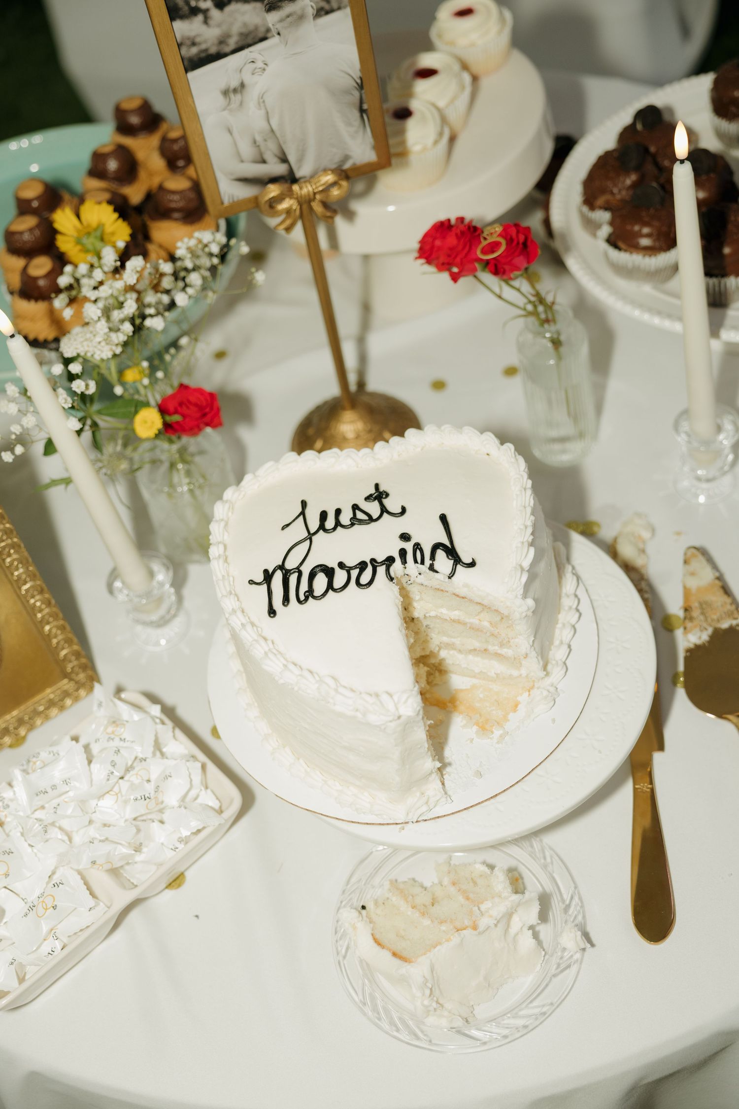 Heart-shaped Just Married wedding cake with a slice removed on a white dessert table.