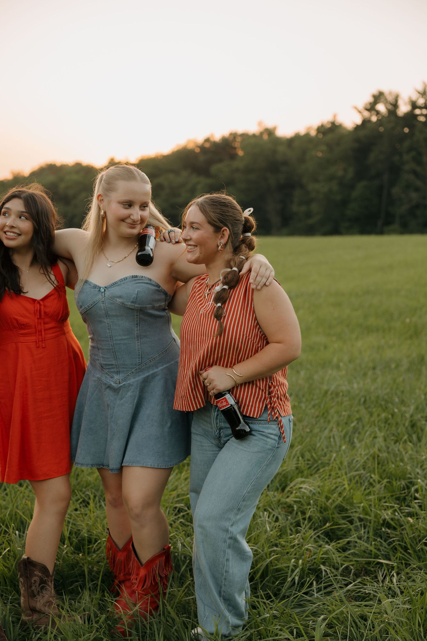Three young women smiling and posing together in a green field at sunset, wearing summer dresses and holding bottles.