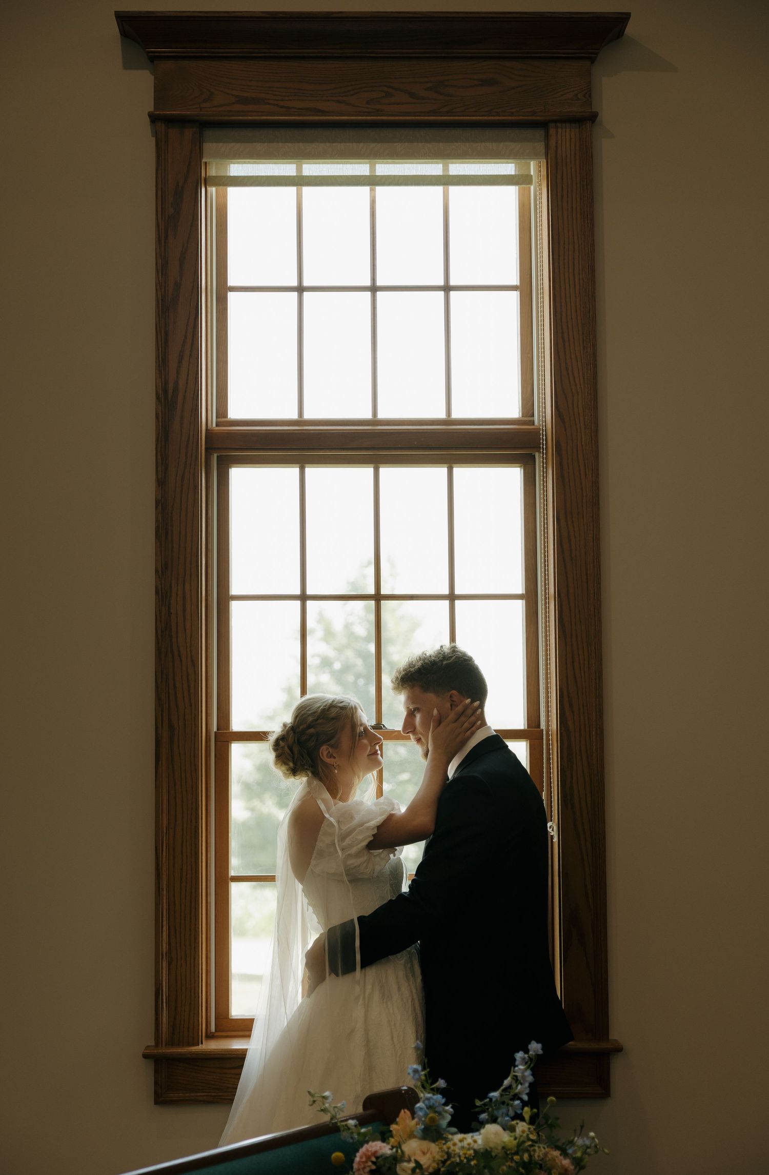 A bride and groom share an intimate moment silhouetted against a tall wooden-framed window on their wedding day.