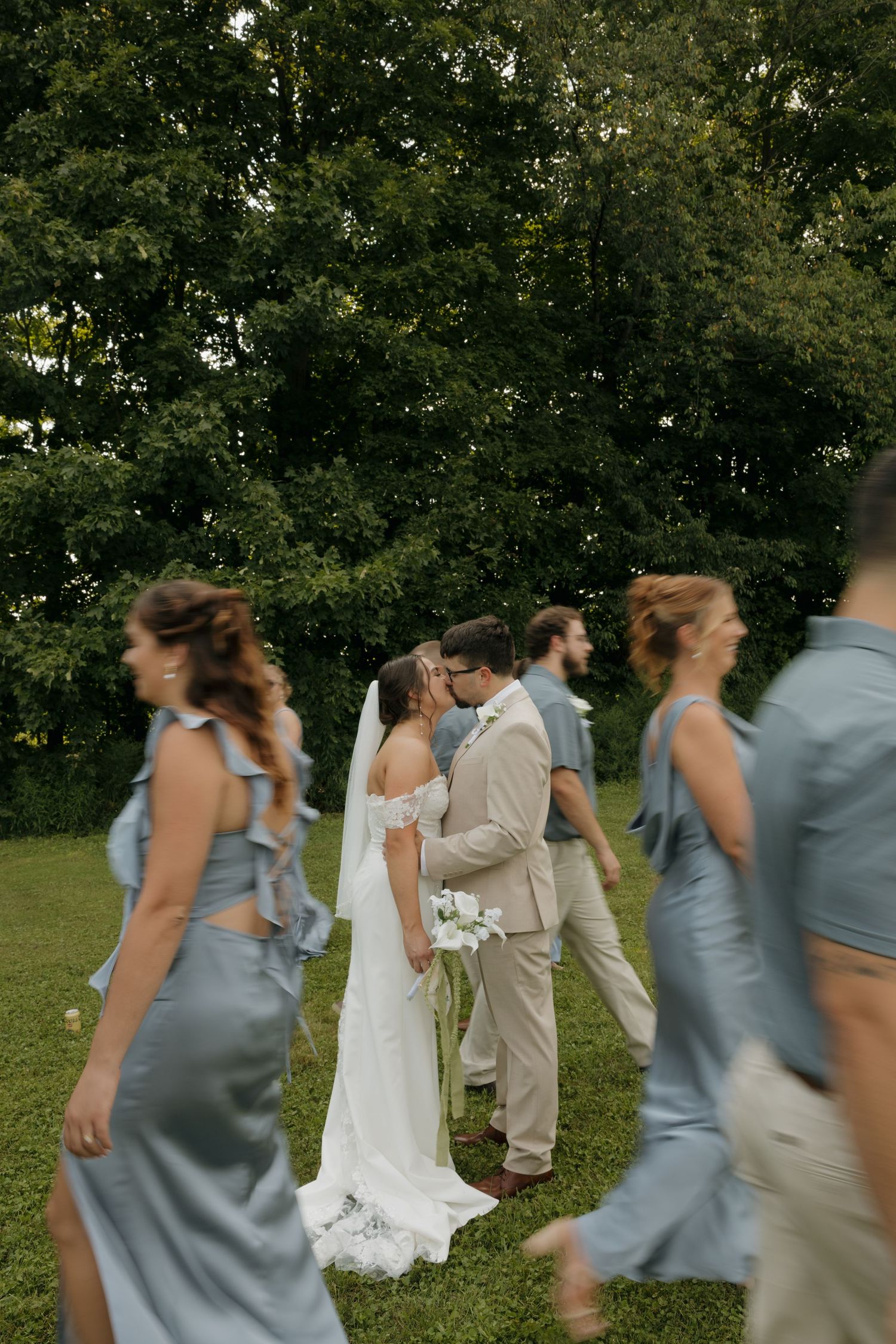 Bride and groom share a kiss while wedding party walks past in dusty blue attire on a lush green lawn.