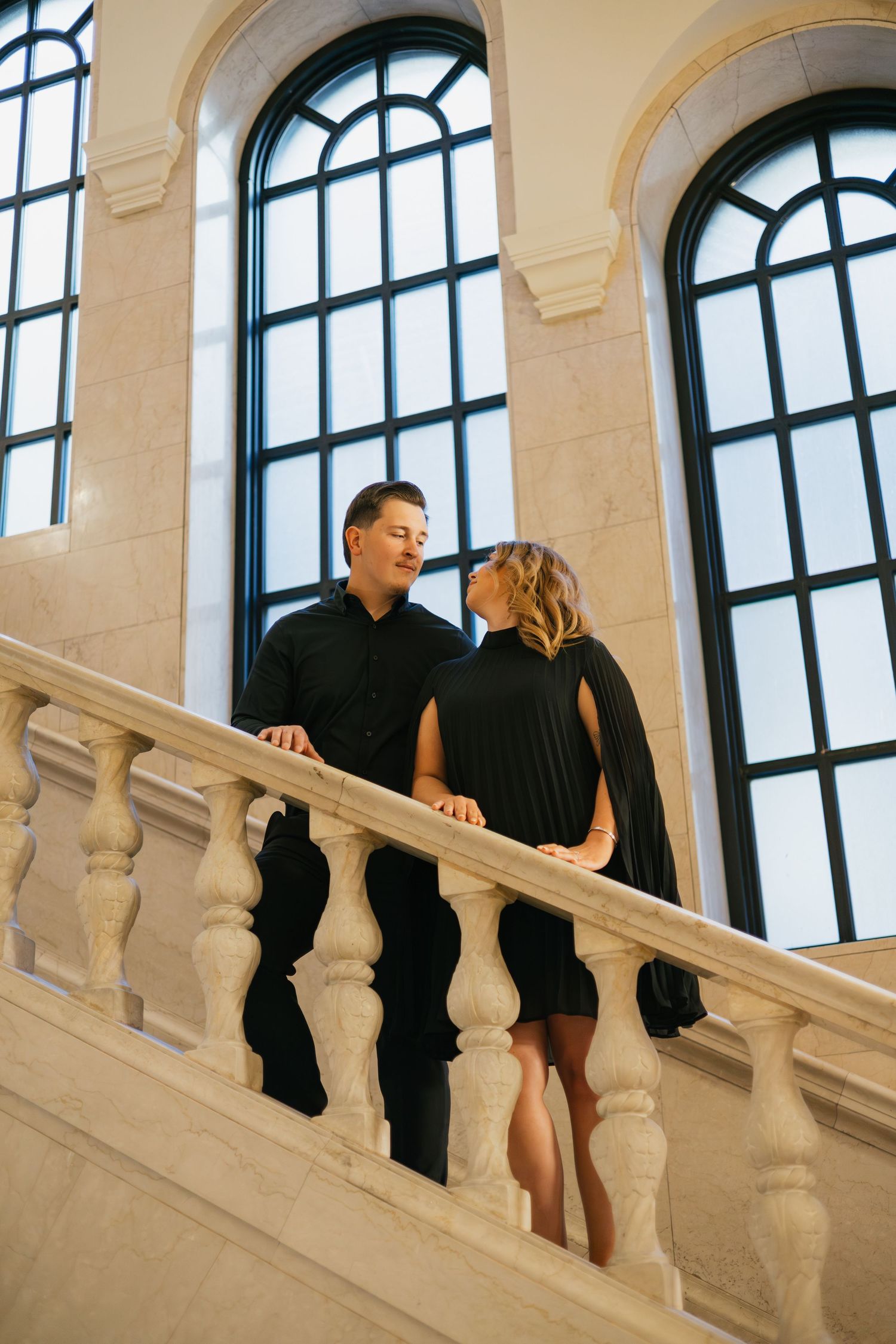 Couple in matching black outfits sharing a romantic moment on an elegant marble staircase with arched windows.