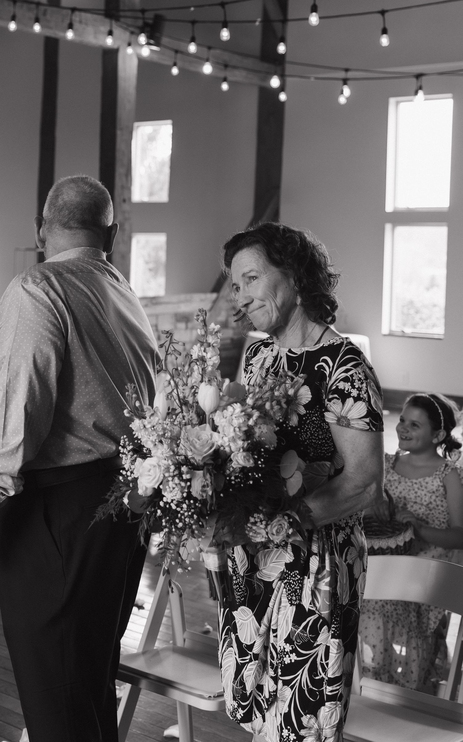 Black and white photo of a woman holding a floral bouquet at an indoor wedding ceremony with string lights overhead.