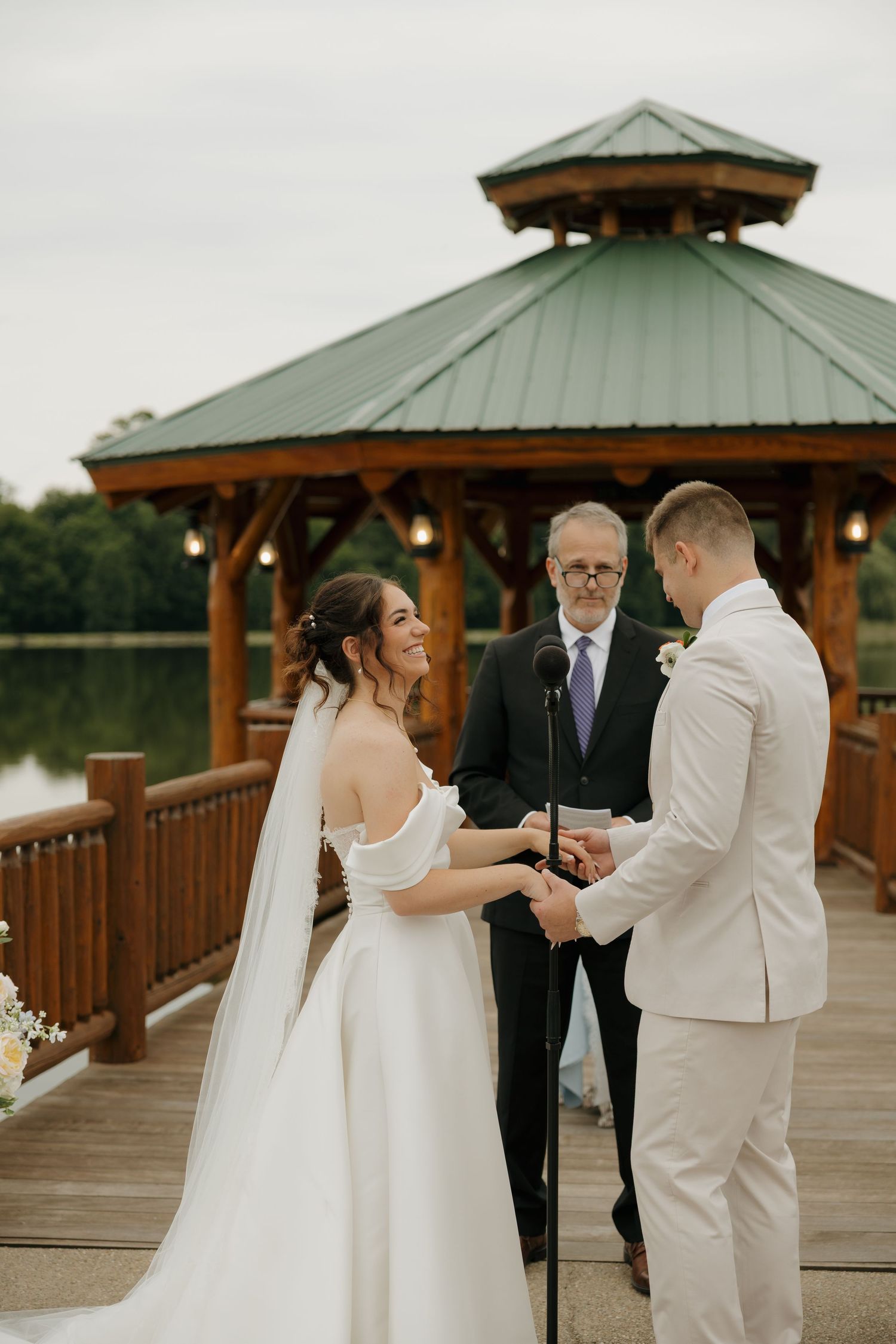 Bride and groom exchange vows on a wooden lakeside deck with a rustic gazebo backdrop during an outdoor wedding ceremony.