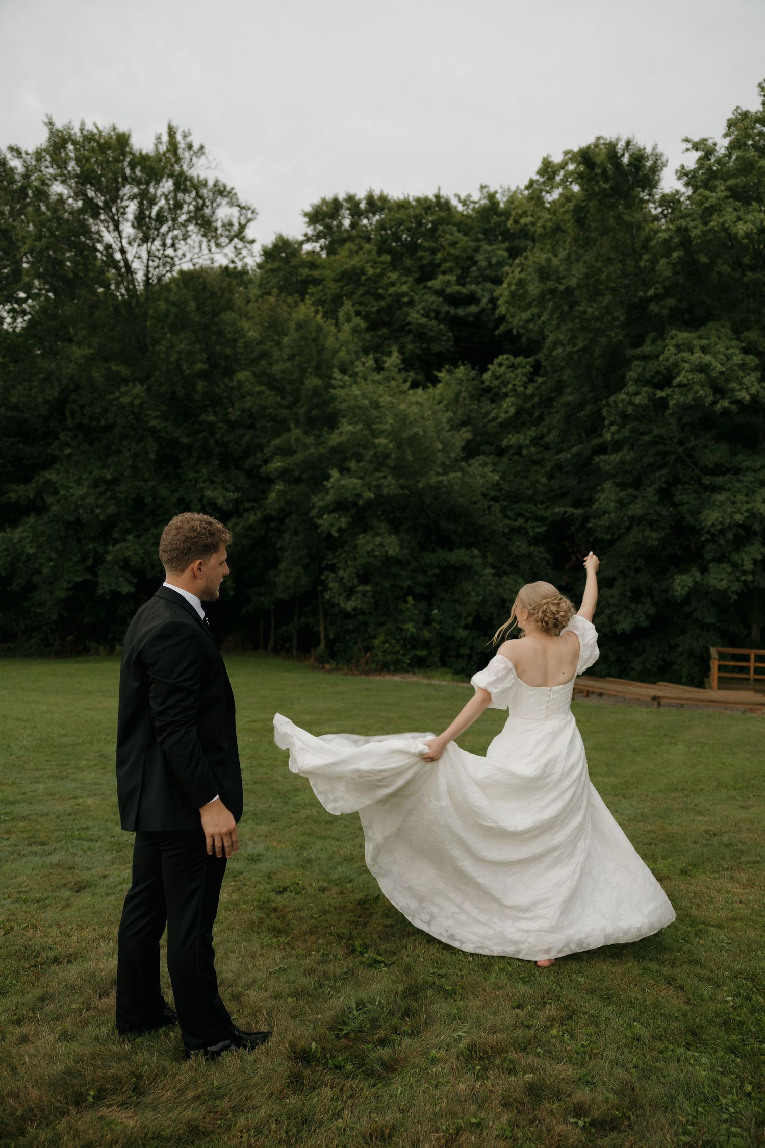 Bride in white gown twirling on green lawn while groom in black suit watches, surrounded by lush trees.