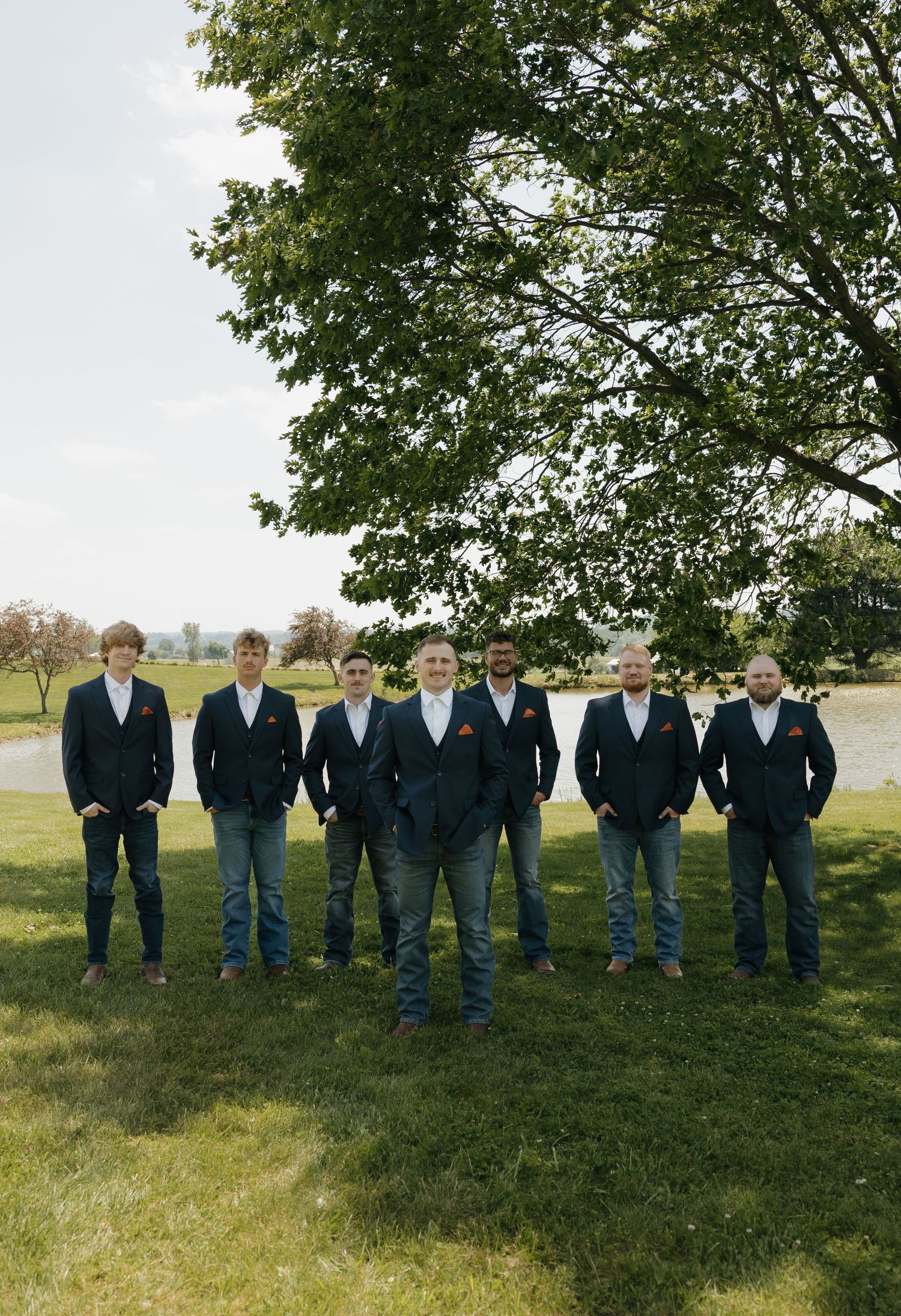 Seven groomsmen in navy blazers and jeans pose outdoors under a large green tree on a sunny wedding day.