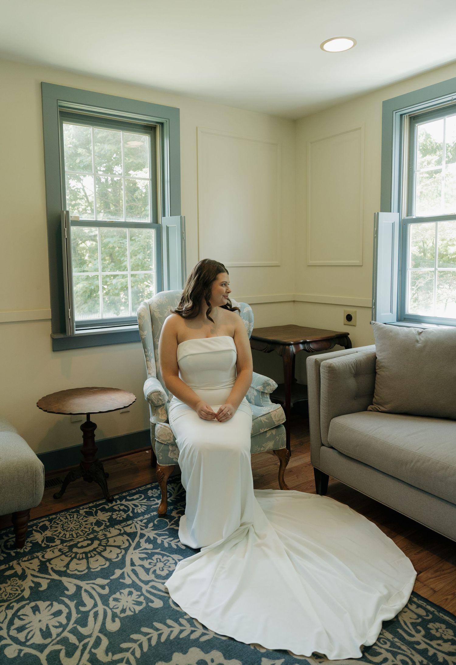 Bride in white strapless gown seated in elegant chair by windows in a cozy room with blue patterned rug.