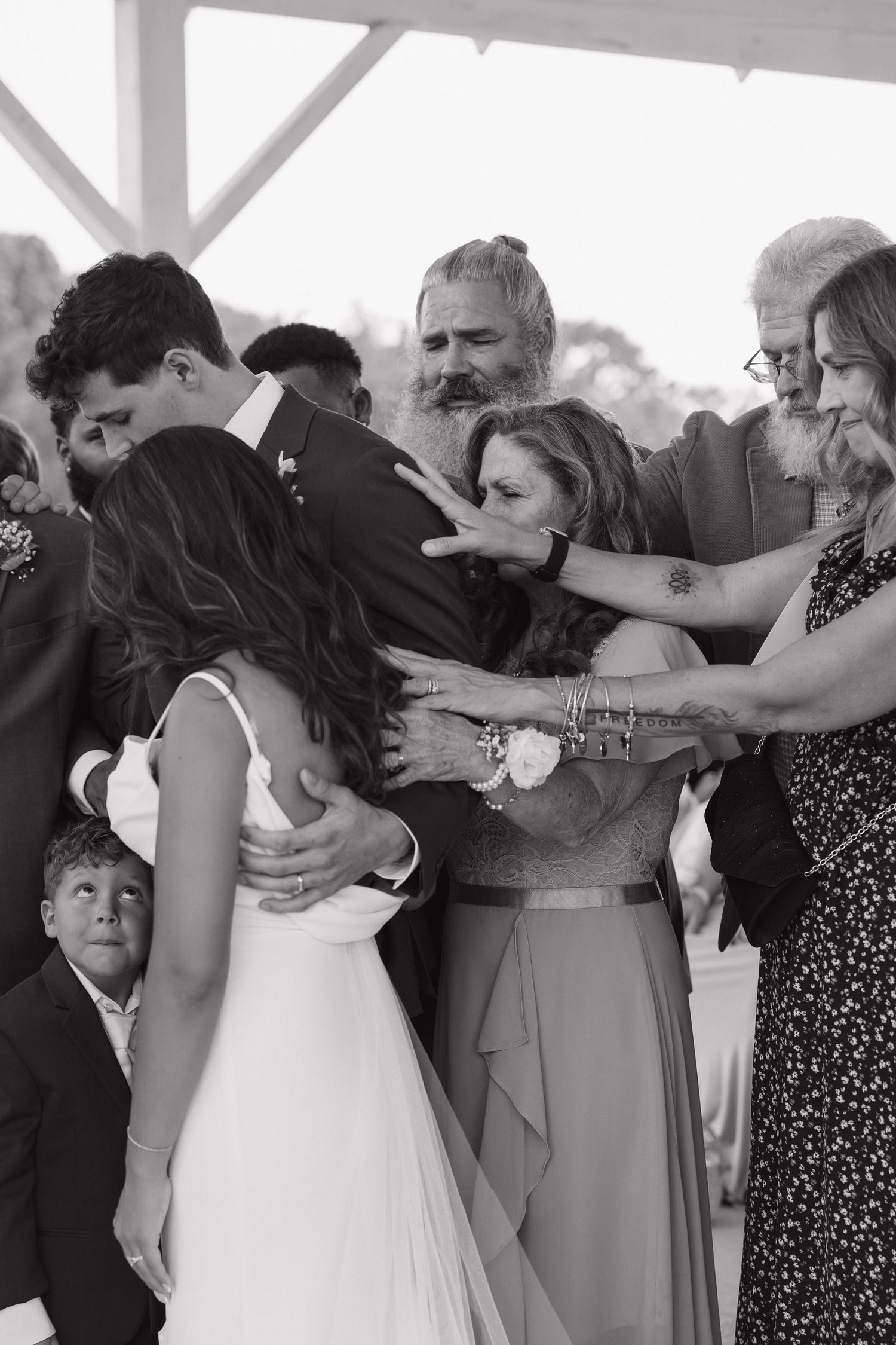 Black and white photo of a bride and groom embraced by emotional family members during a wedding ceremony.