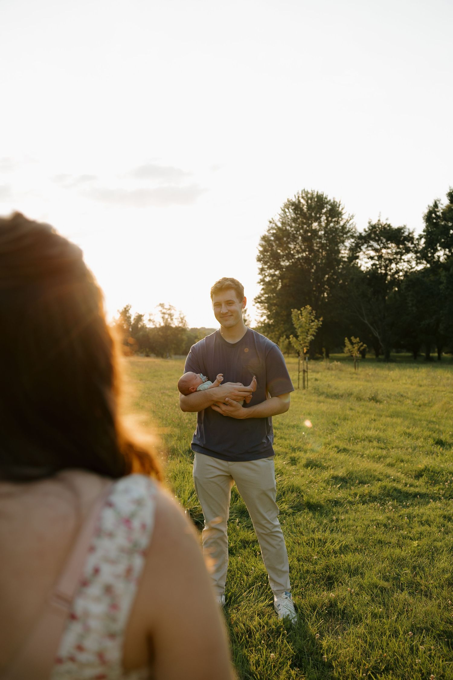 A man holds a newborn baby in a sunlit green field at golden hour, smiling at a woman in the foreground.