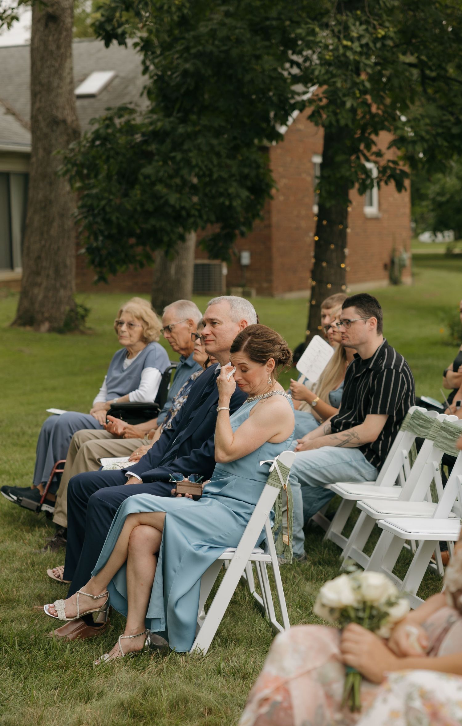 Wedding guests seated on white chairs outdoors, emotional woman in blue dress wiping tears during ceremony.