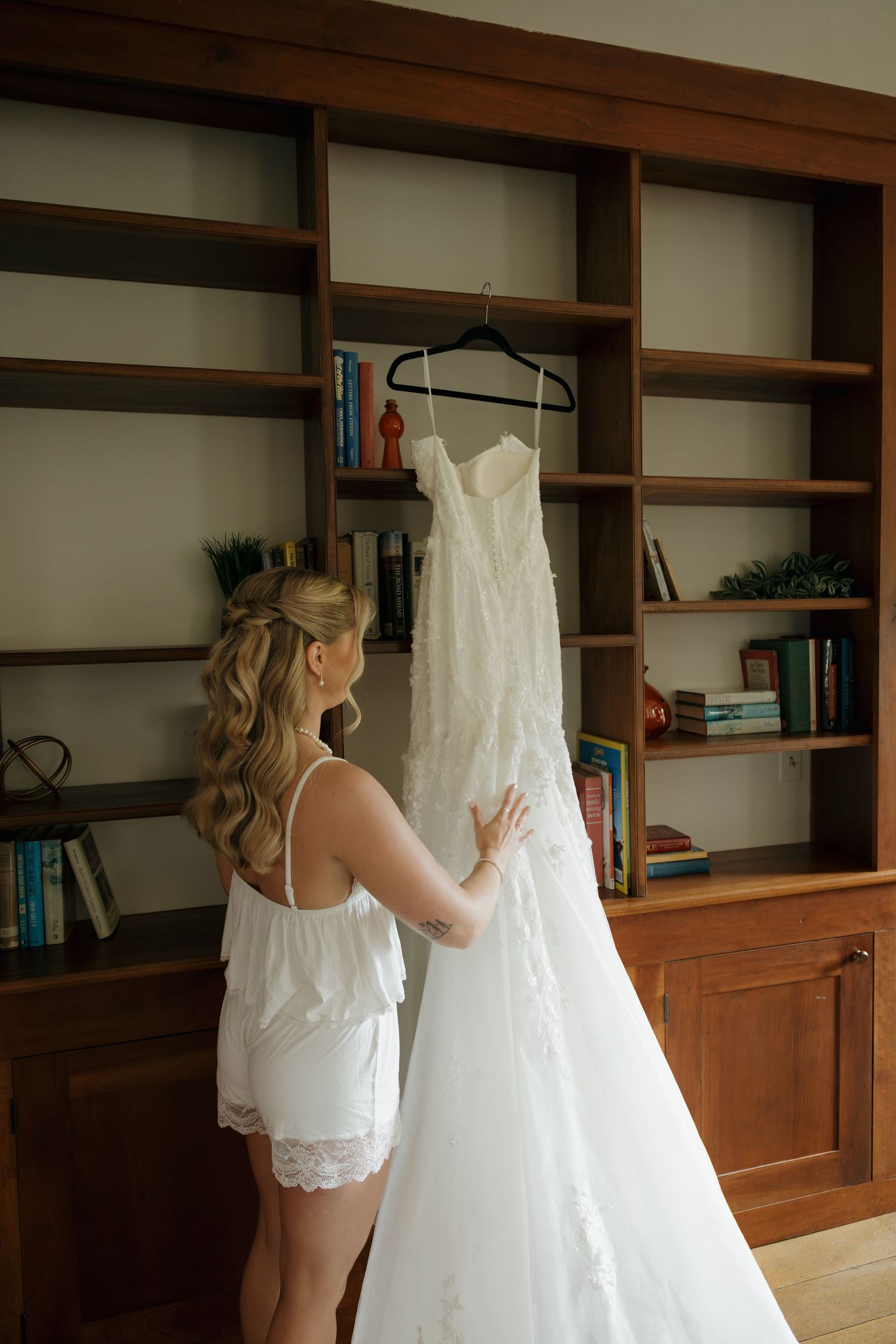 Bride in white lace robe admiring her wedding gown hanging on a bookshelf in a warmly lit room.