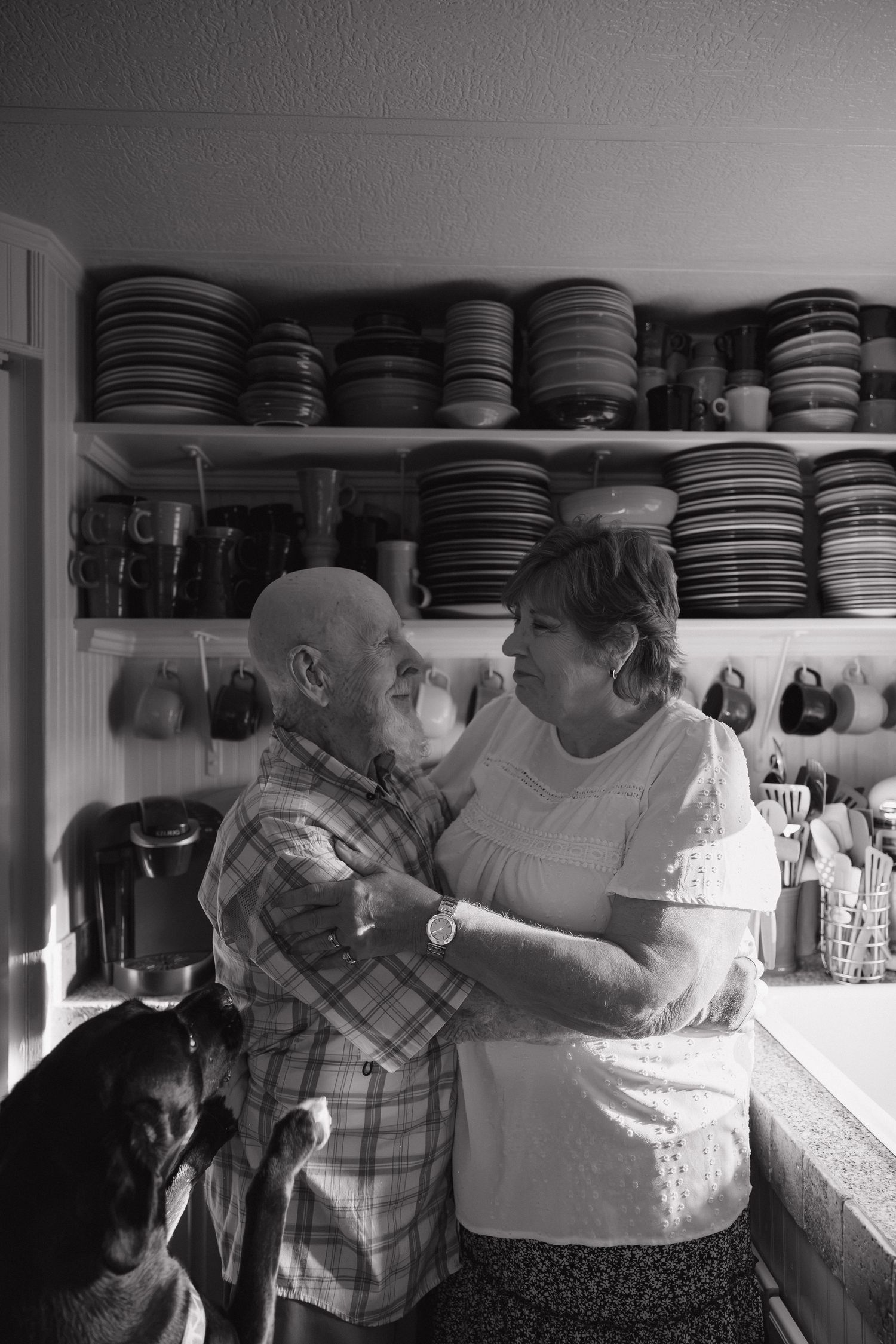 Elderly couple sharing a tender moment dancing in a kitchen with shelves of dishes in black and white photo.