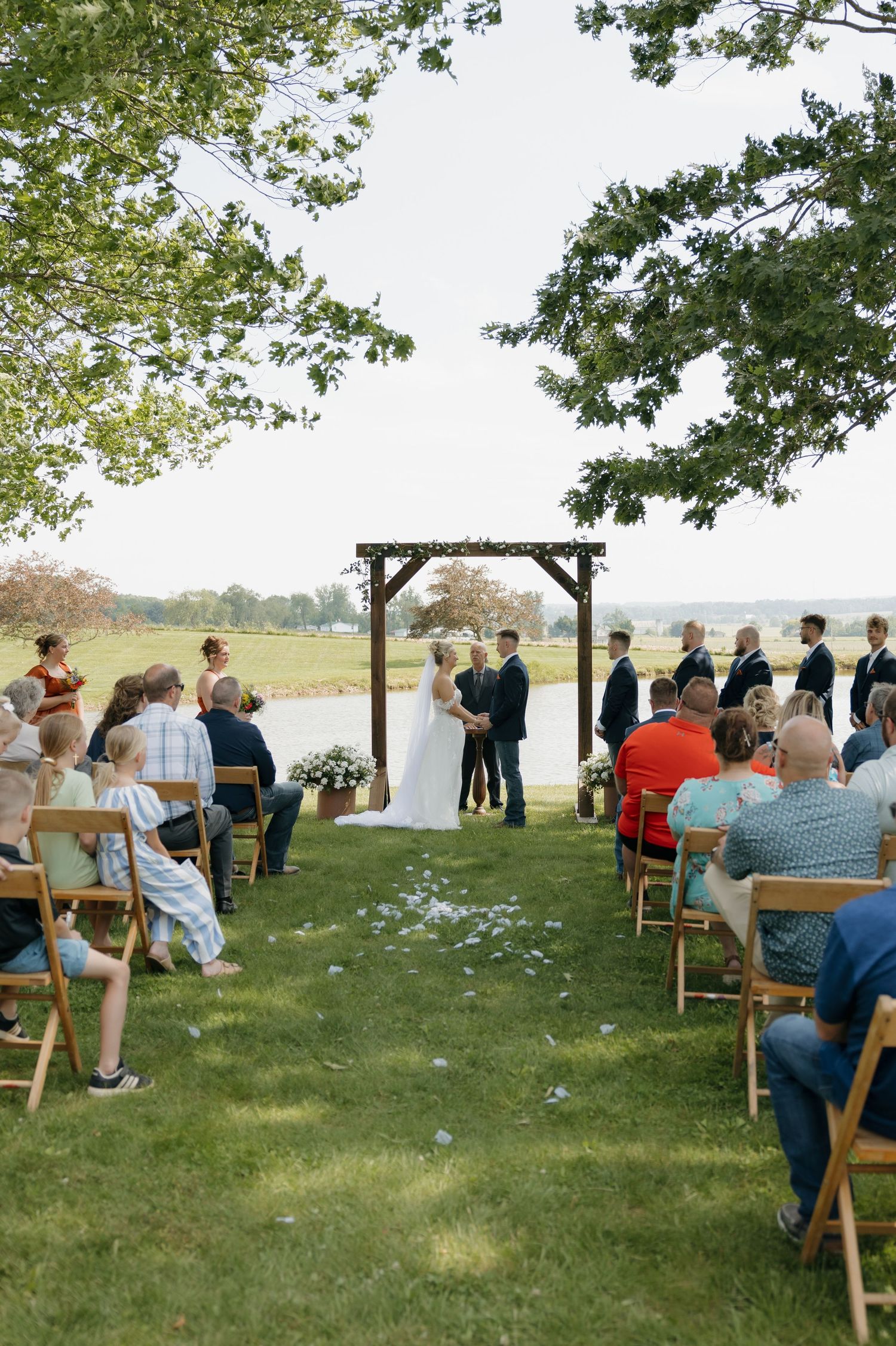 Outdoor wedding ceremony on green lawn with wooden arch, bride and groom, and seated guests under large trees.