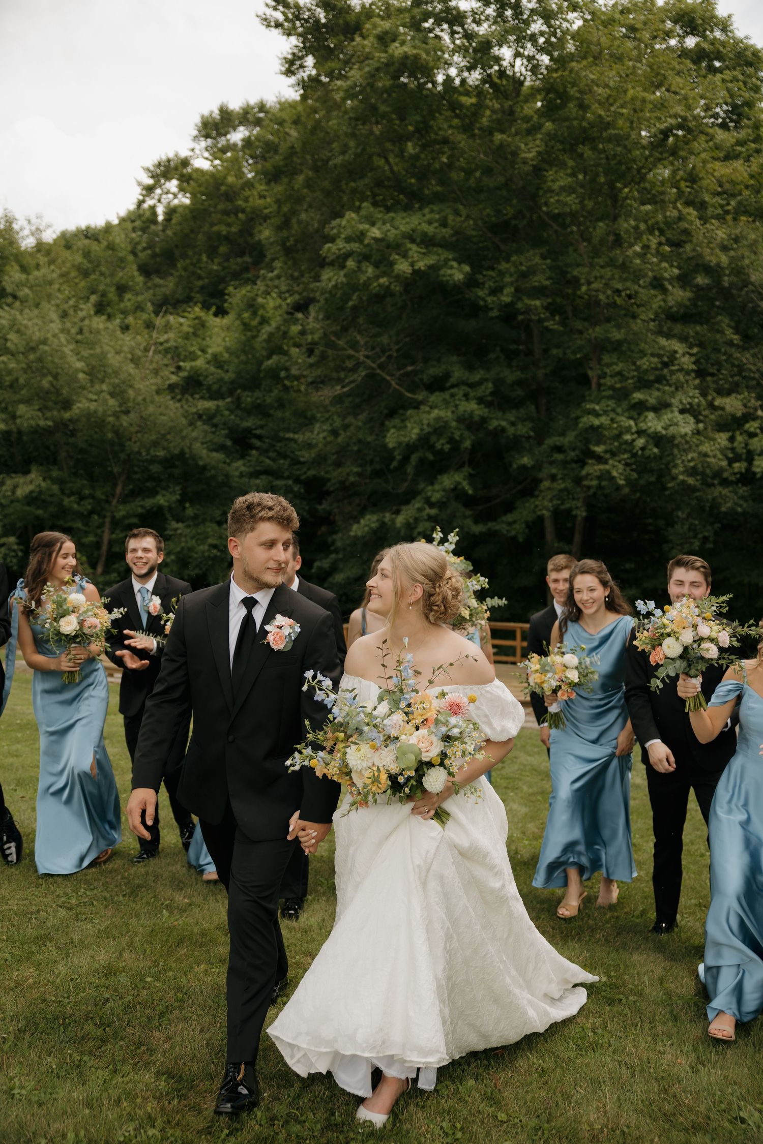Bride and groom walk hand-in-hand outdoors, followed by wedding party in blue gowns and black suits, with lush green trees behind.