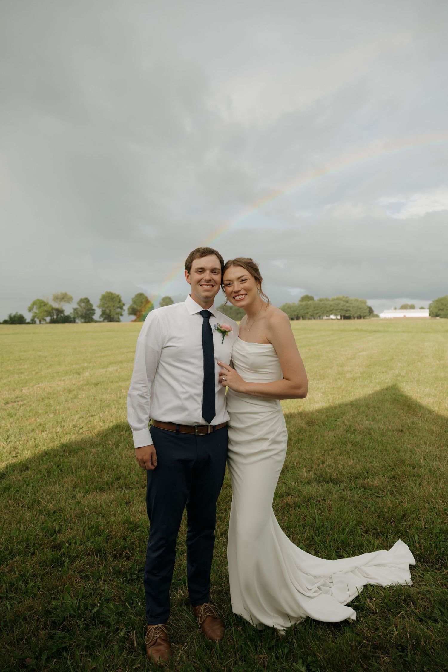 Bride and groom smiling in a green field with a rainbow in the cloudy sky behind them on their wedding day.