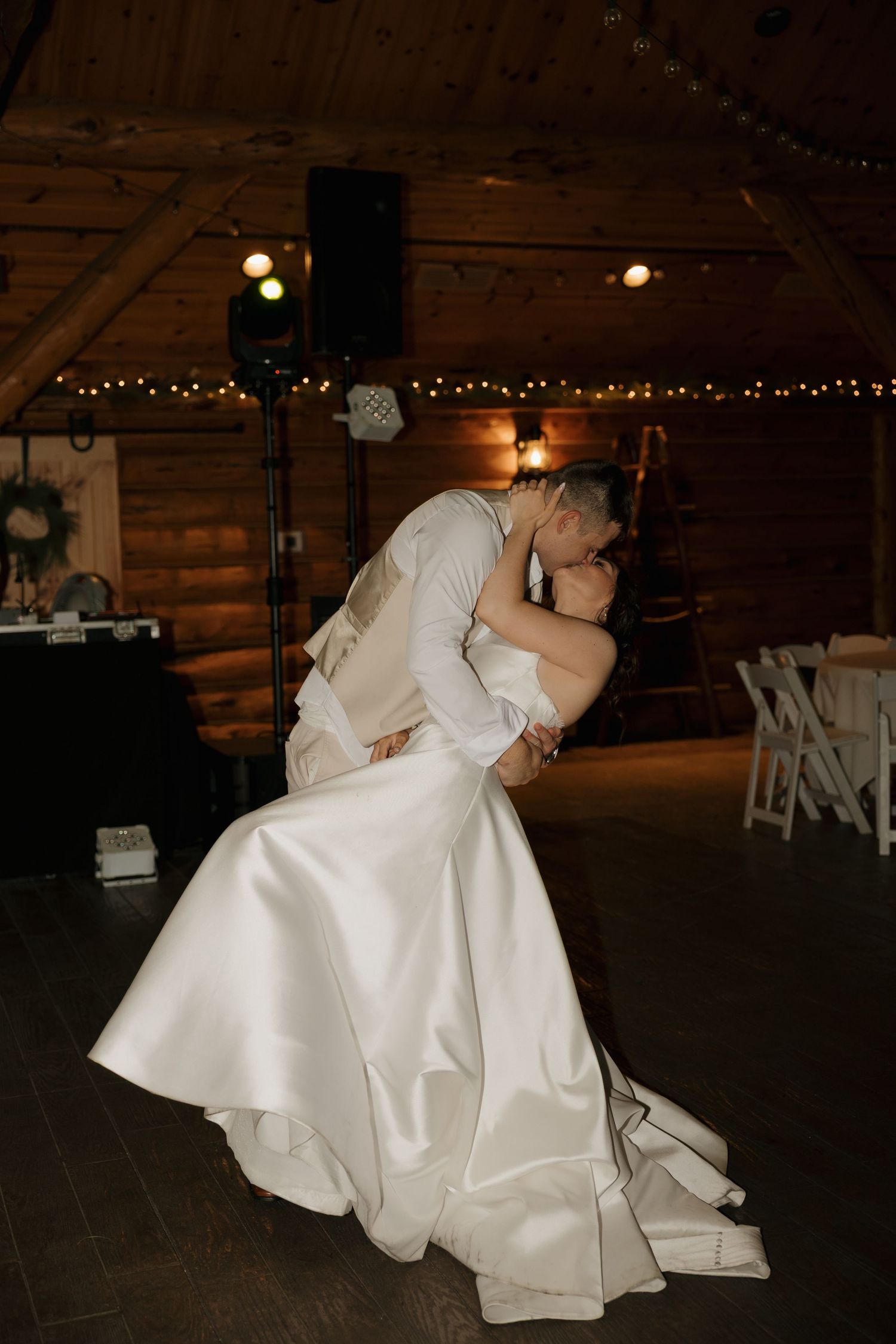 Bride and groom share a romantic dip kiss on the dance floor of a rustic log cabin wedding venue with string lights.