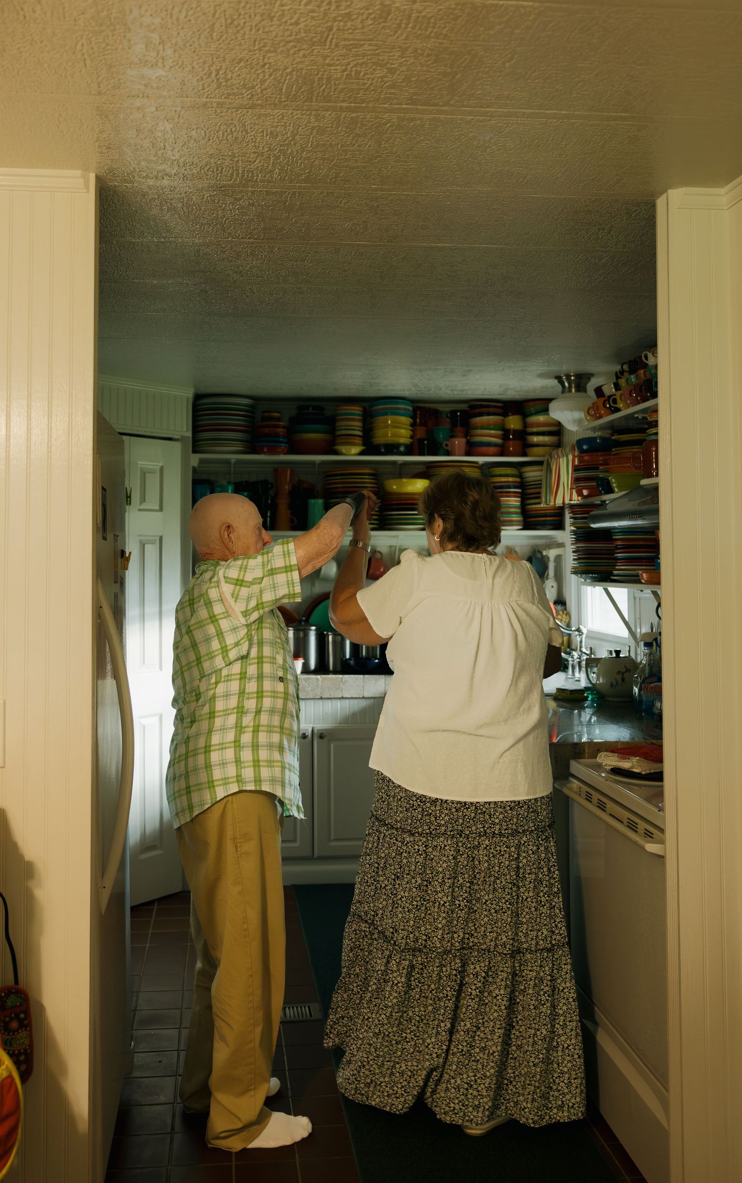 Elderly couple reaching up to shelves filled with colorful plates and dishes in a cozy kitchen.