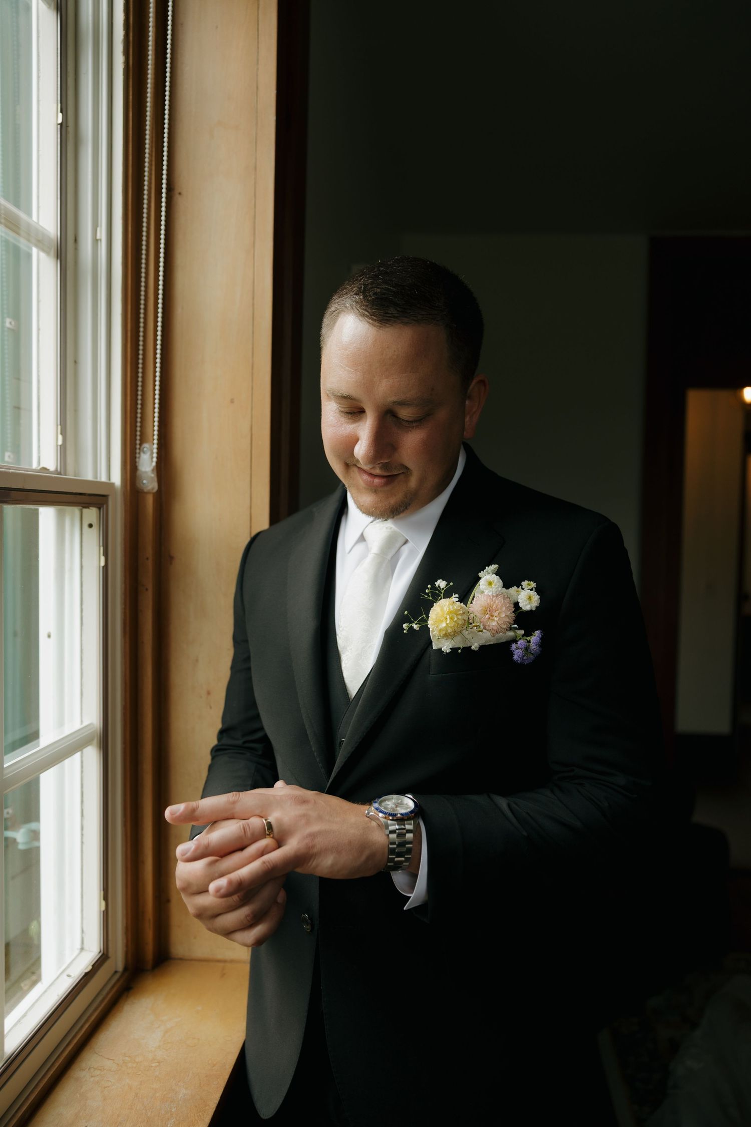 Groom in black tuxedo with floral boutonniere adjusts cufflinks by window on wedding day.