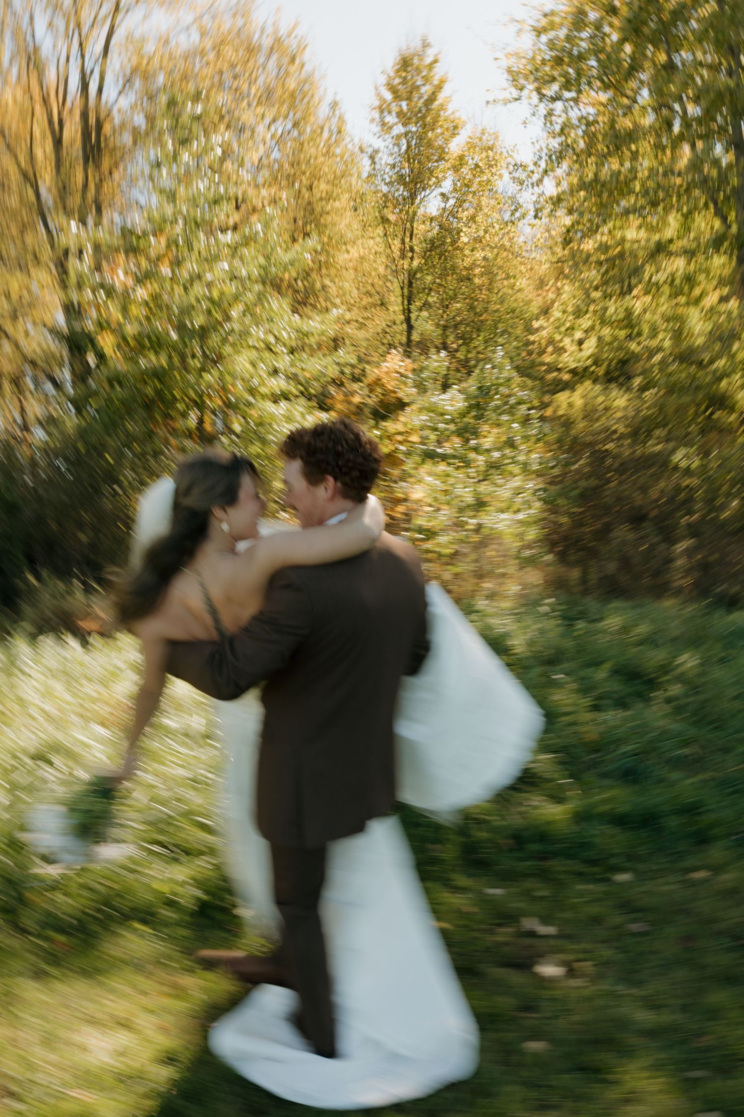 Motion-blurred bride in a white gown embracing a groom in a brown suit while twirling on a grassy path surrounded by colorful autumn trees.