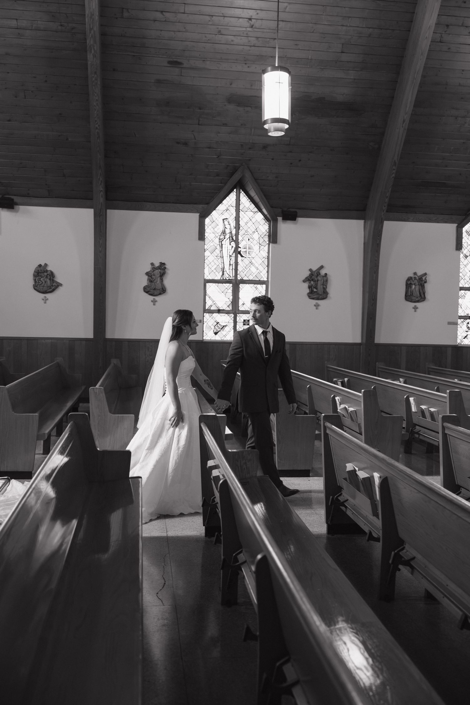 Black and white photo of a bride in a veil and groom in a suit holding hands between wooden pews in a church.