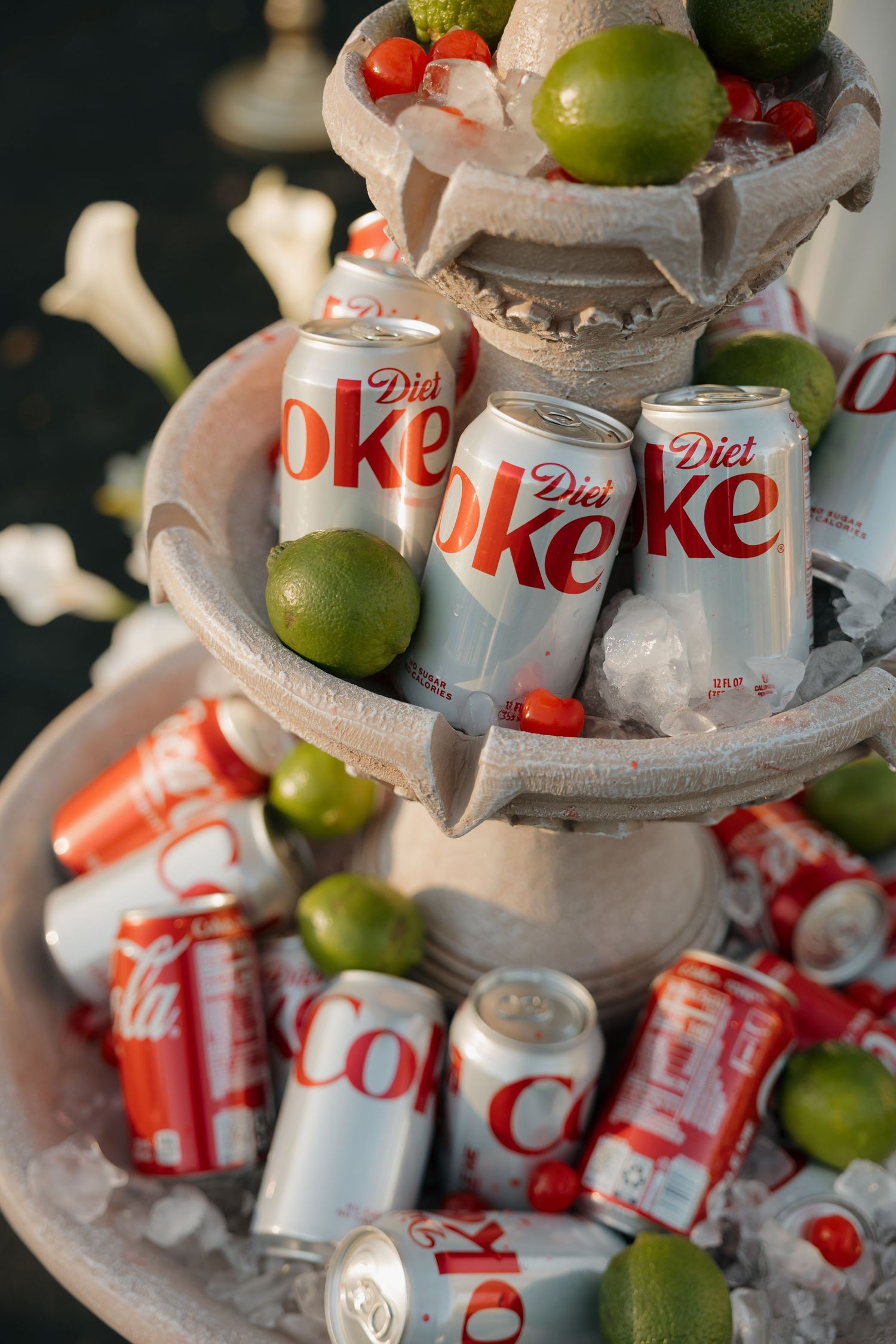 Diet Coke and Coca-Cola cans chilled on ice with fresh limes and cherries displayed in a tiered stone fountain.