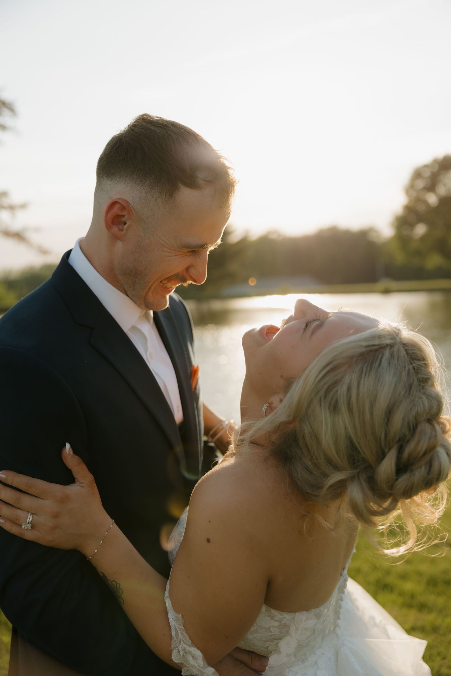 Bride and groom share a joyful moment at golden hour by a lake, bride laughing in a braided updo and white gown.
