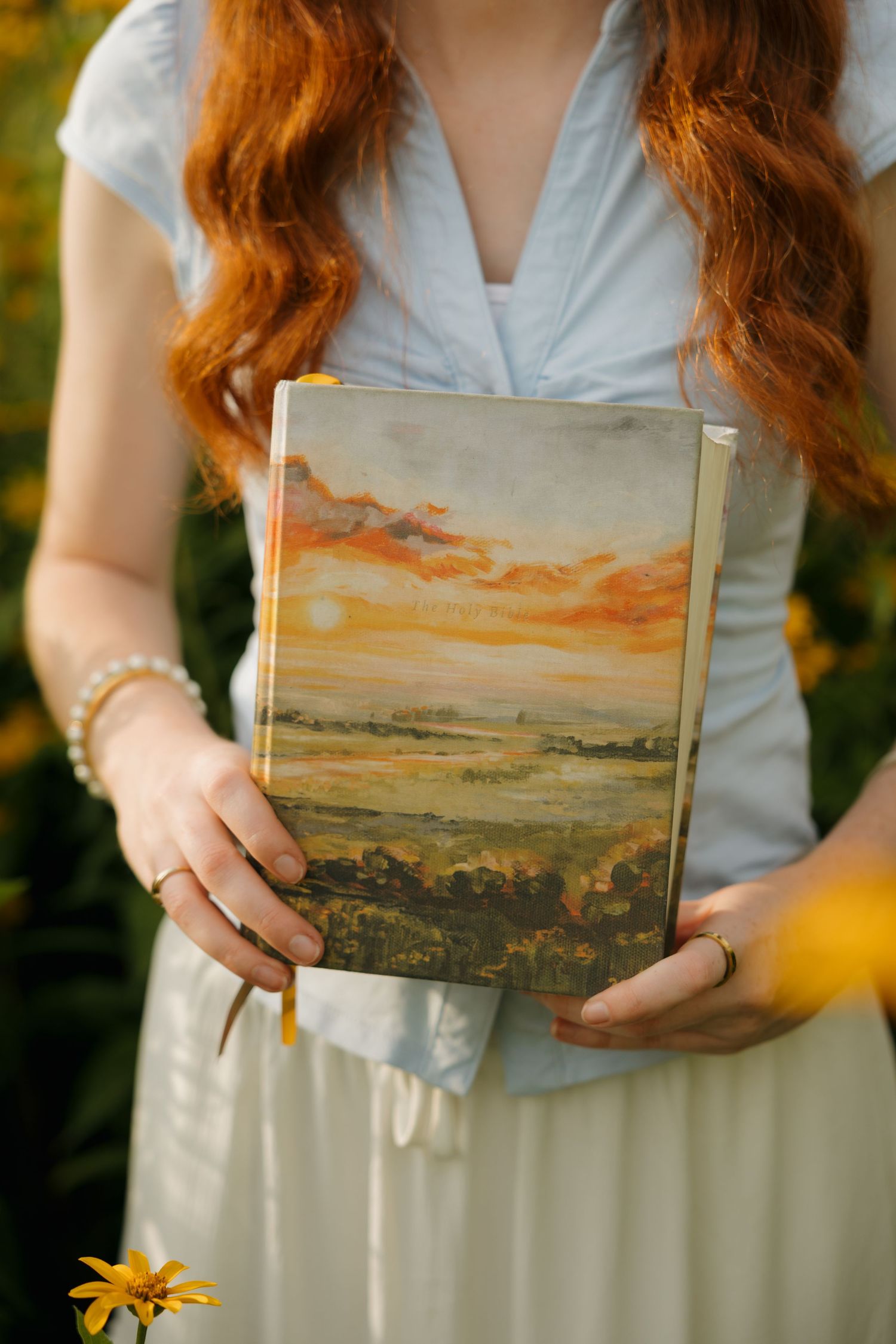 Red-haired woman in white dress holding a book with a painted sunset landscape cover outdoors.