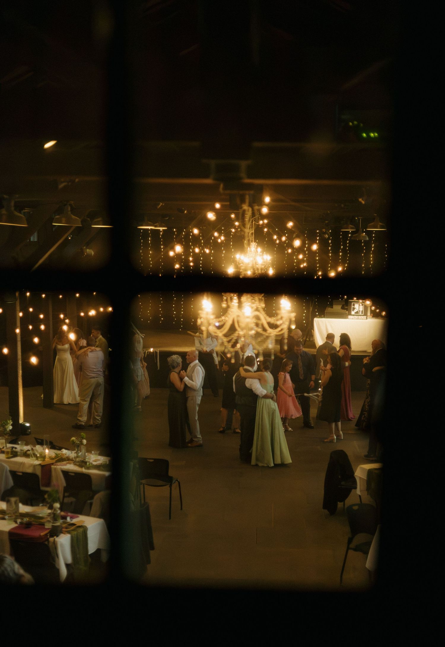 Elegant wedding reception viewed through a window, with guests dancing under chandeliers and string lights in a warmly lit venue.