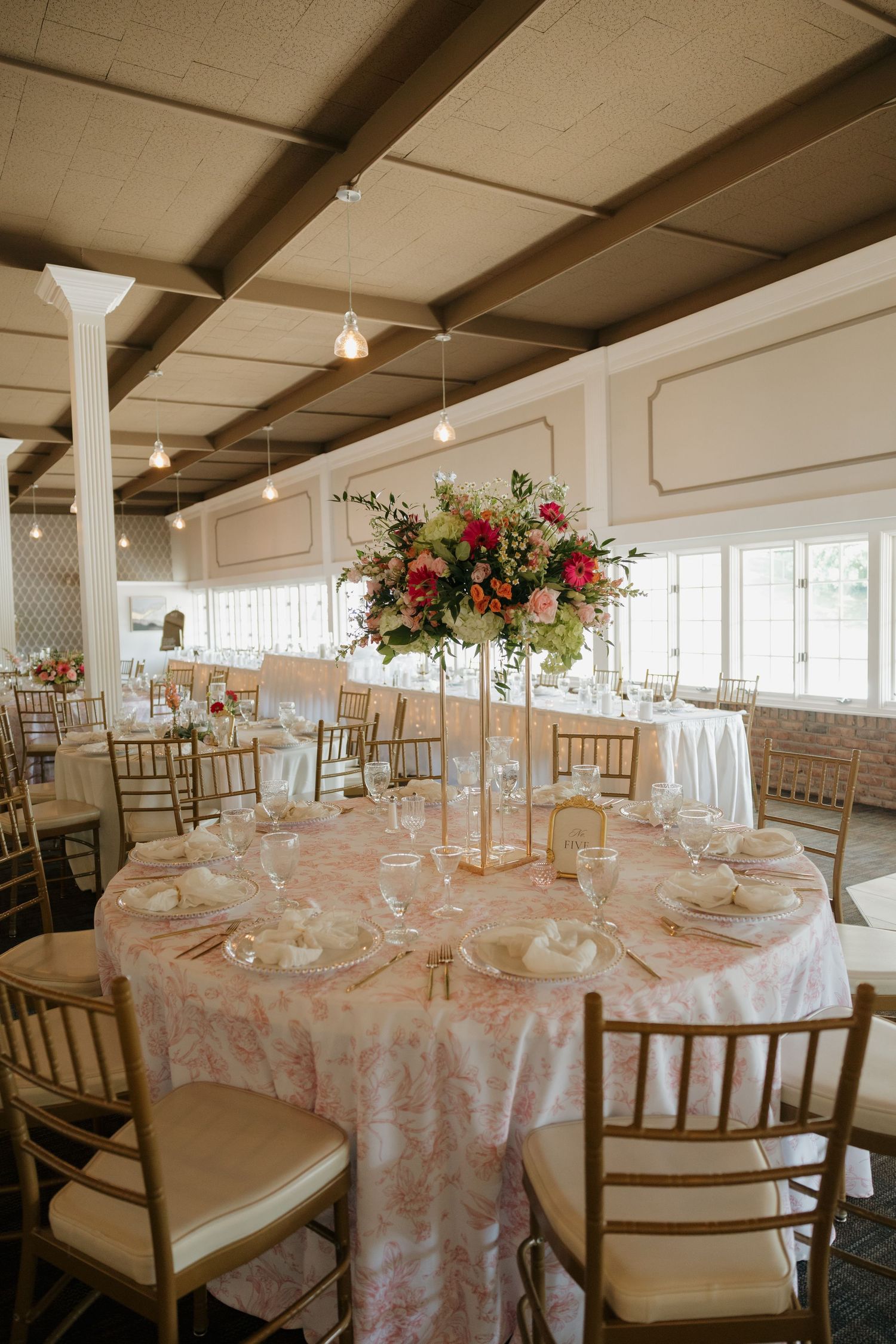 Elegant wedding reception hall with floral centerpieces, gold chiavari chairs, and pink patterned tablecloths under pendant lights.
