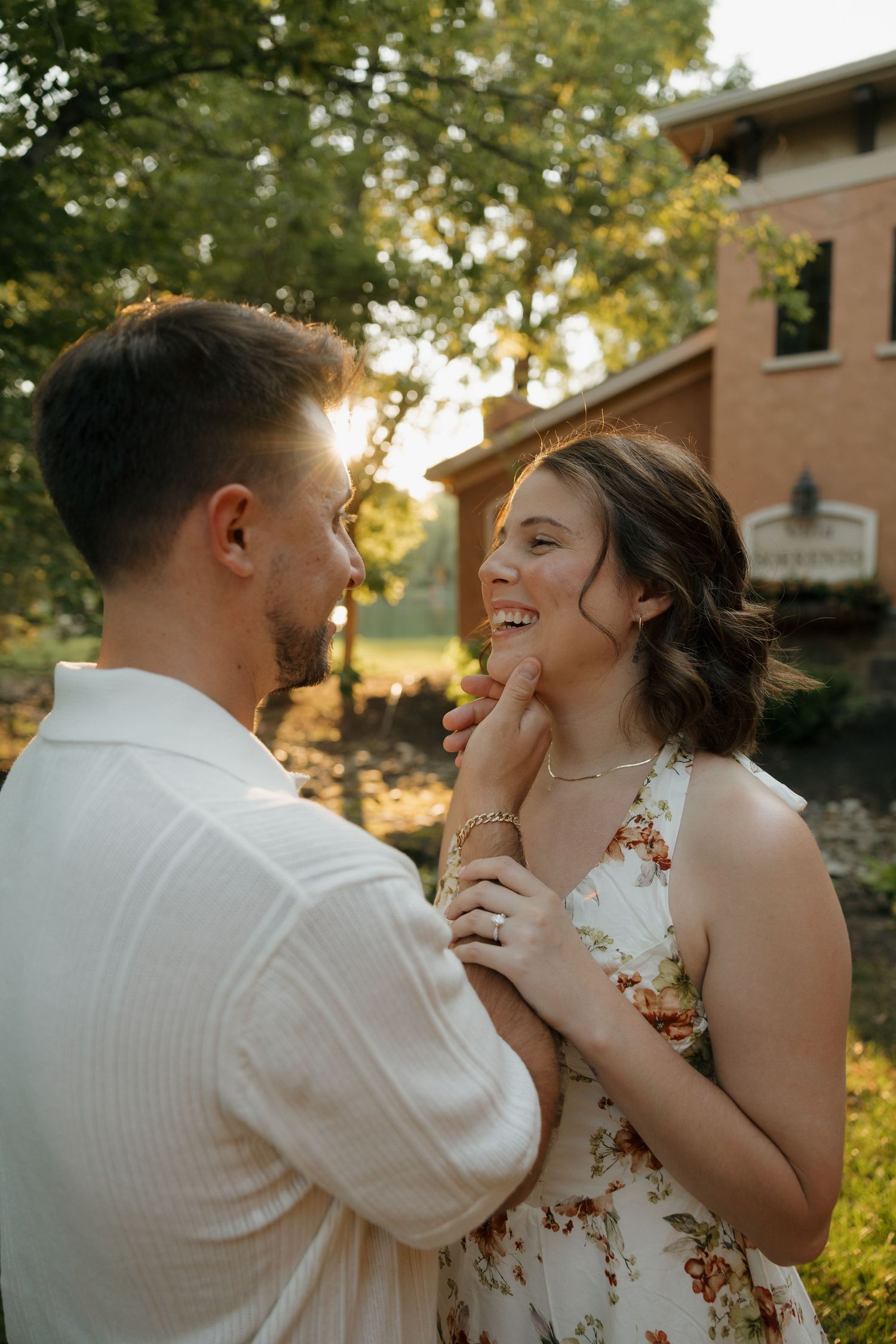 Couple sharing a tender moment outdoors at golden hour, woman in floral dress laughing as man gently holds her face.