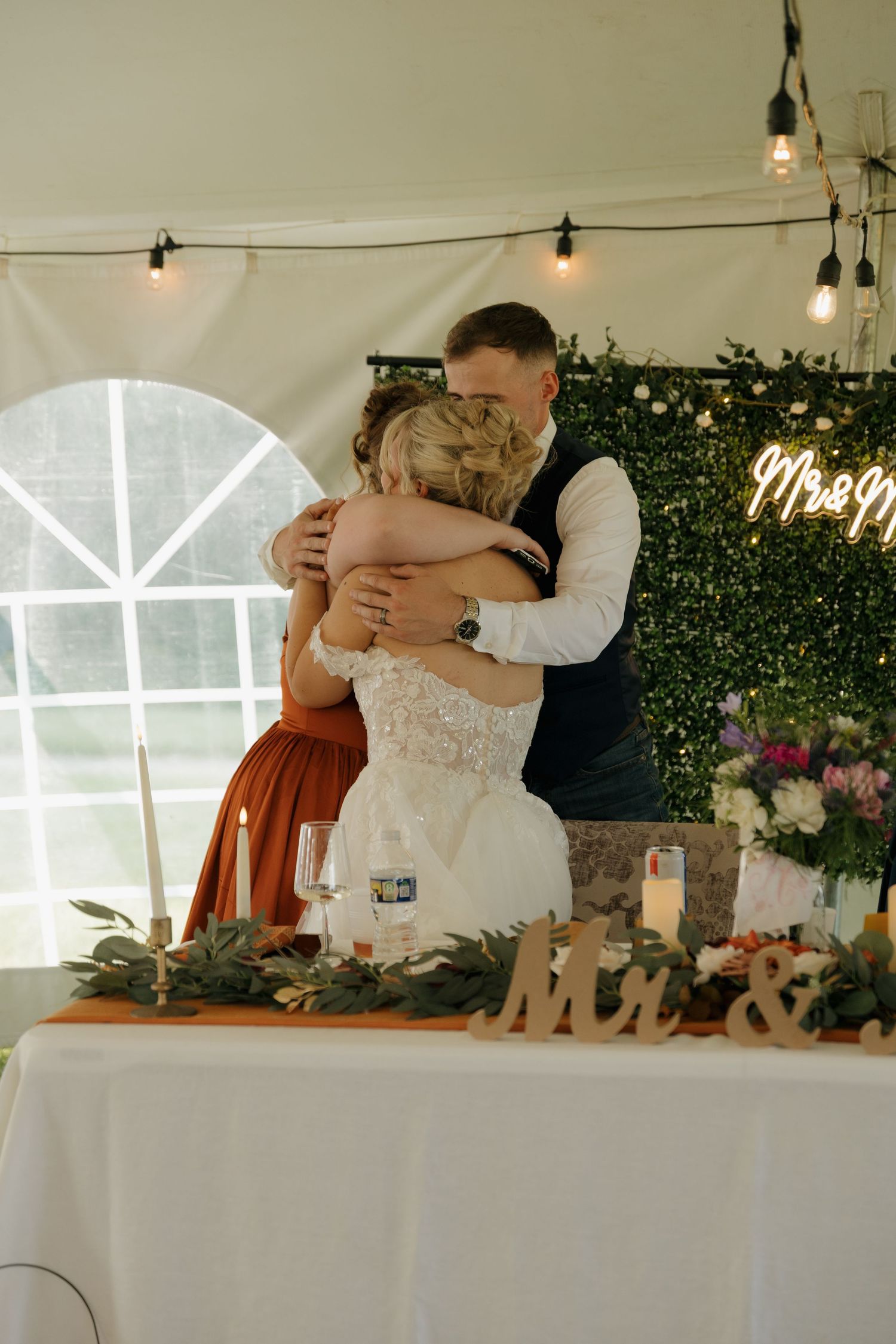 Bride and groom share a tender hug at their wedding reception sweetheart table decorated with greenery and Mr & Mrs sign.