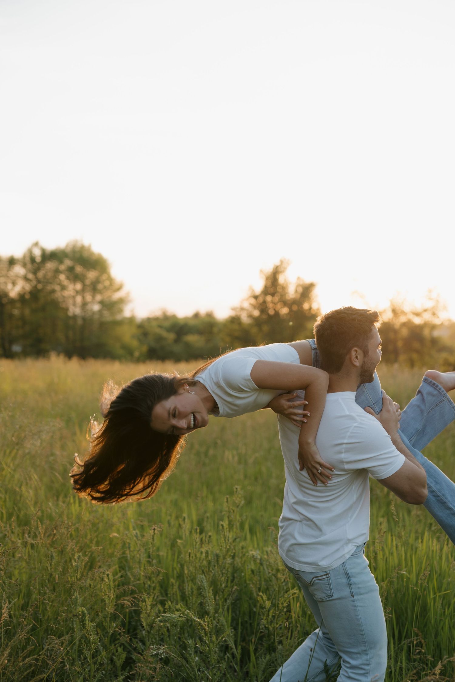 A couple playfully dancing in a sunlit green field at sunset, the woman leaning back joyfully in her partner's arms.