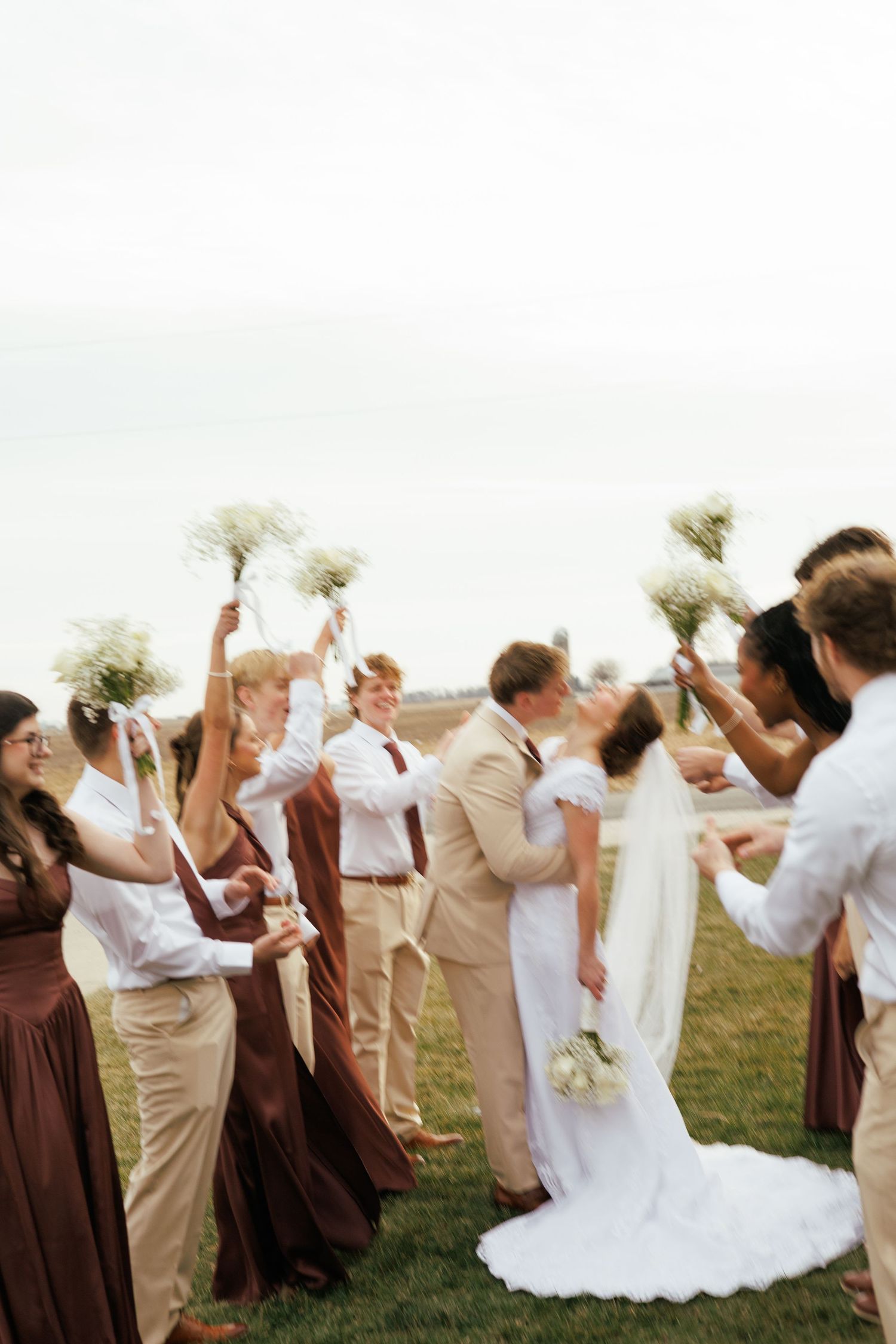 Wedding party in brown and white attire celebrates with joyful poses in an outdoor field.