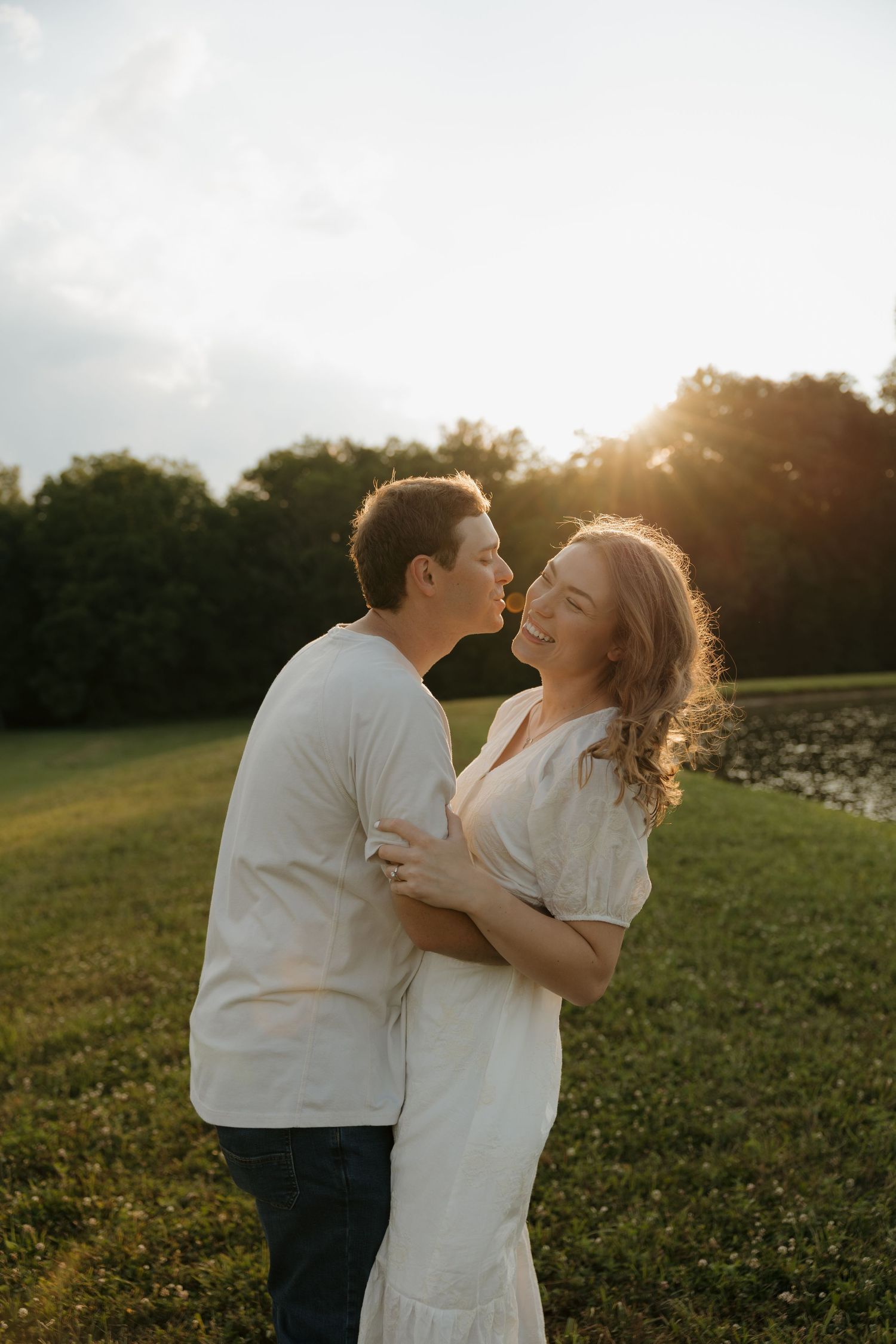 Couple sharing a romantic kiss at golden hour in a green field with warm sunset light glowing behind them.