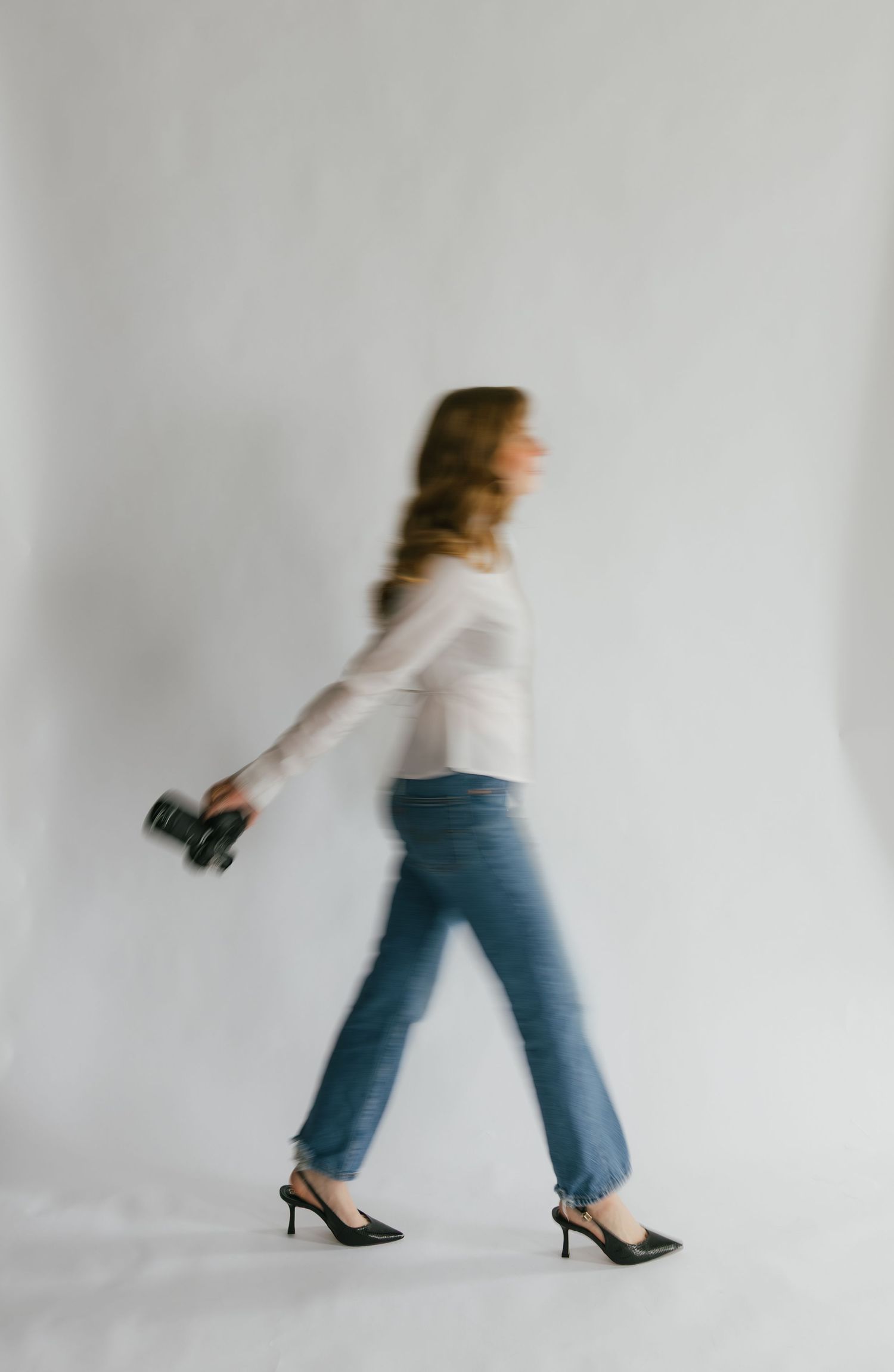 A woman walks in motion blur wearing jeans, white top, and heels while holding a camera against a white backdrop.